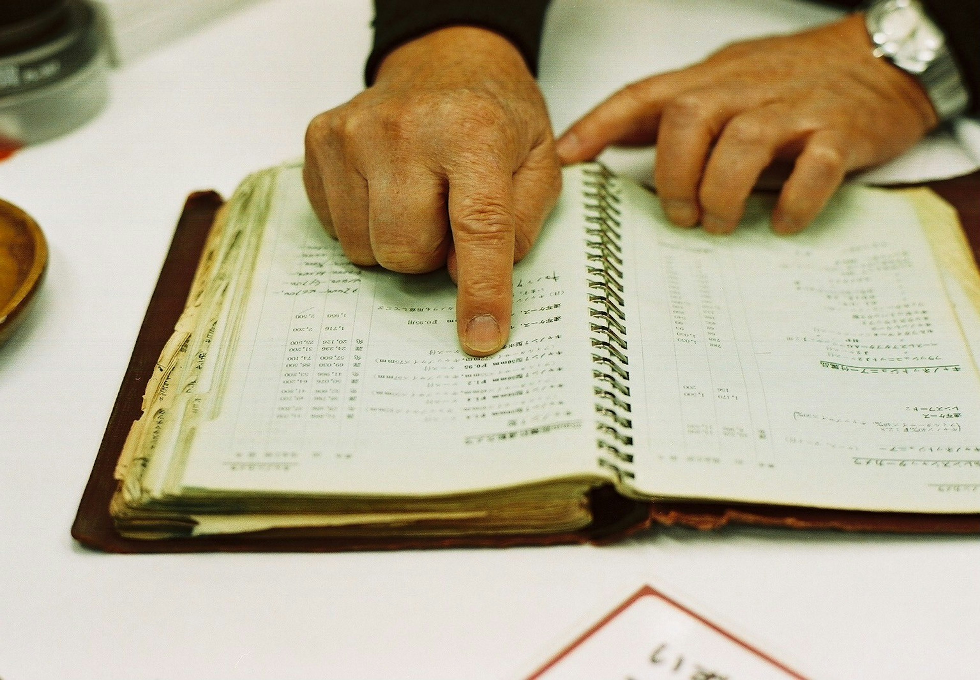 Una mano señalando un cuaderno antiguo con notas manuscritas y números visibles