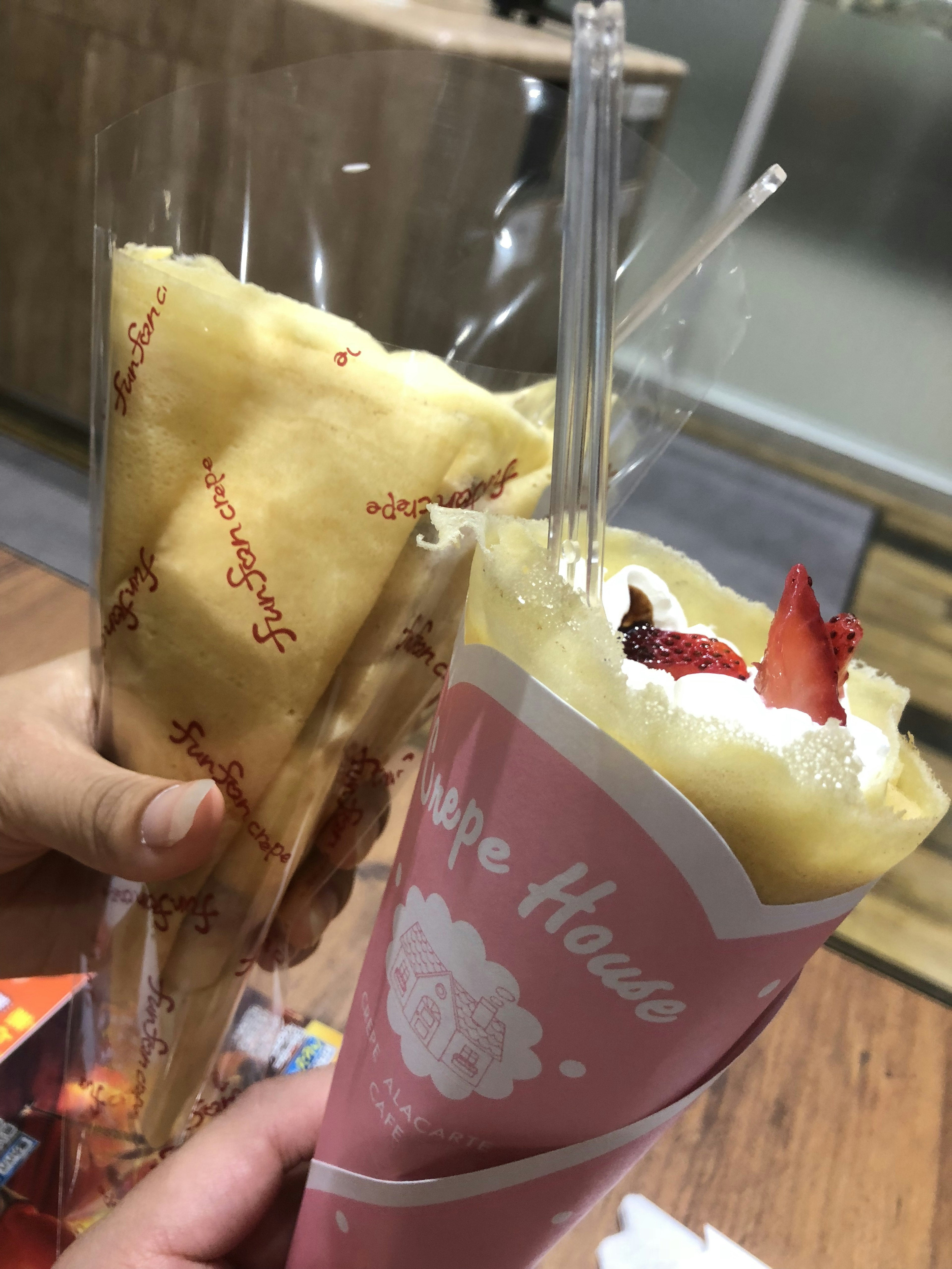 二つのクレープが手に持たれています 一つはストロベリークリームトッピングのピンクのパッケージ もう一つはシンプルなクリームの黄色いパッケージ