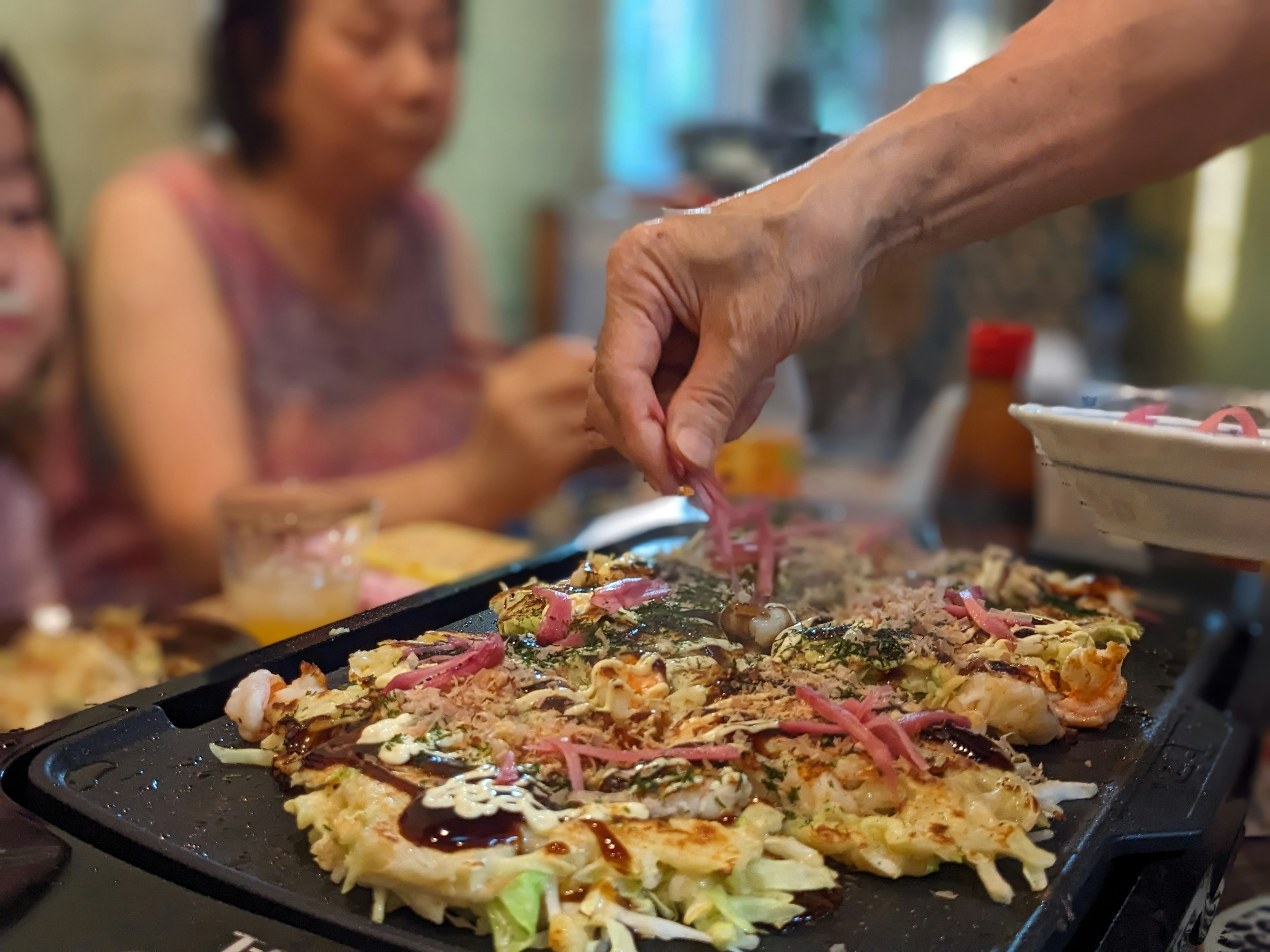 Kumpul keluarga di sekitar plat panas memasak okonomiyaki dengan tangan yang menambahkan topping