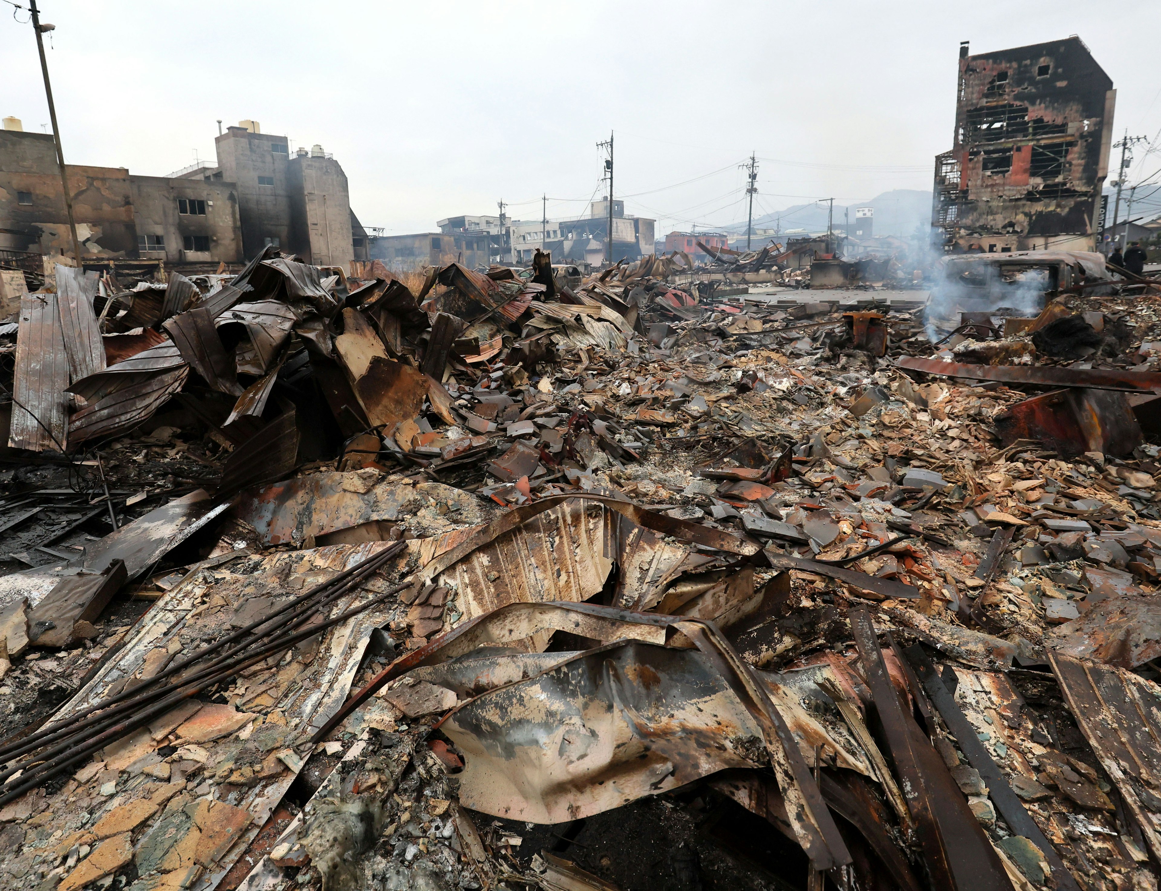 Paisaje devastado de escombros quemados con humo elevándose y edificios en ruinas al fondo