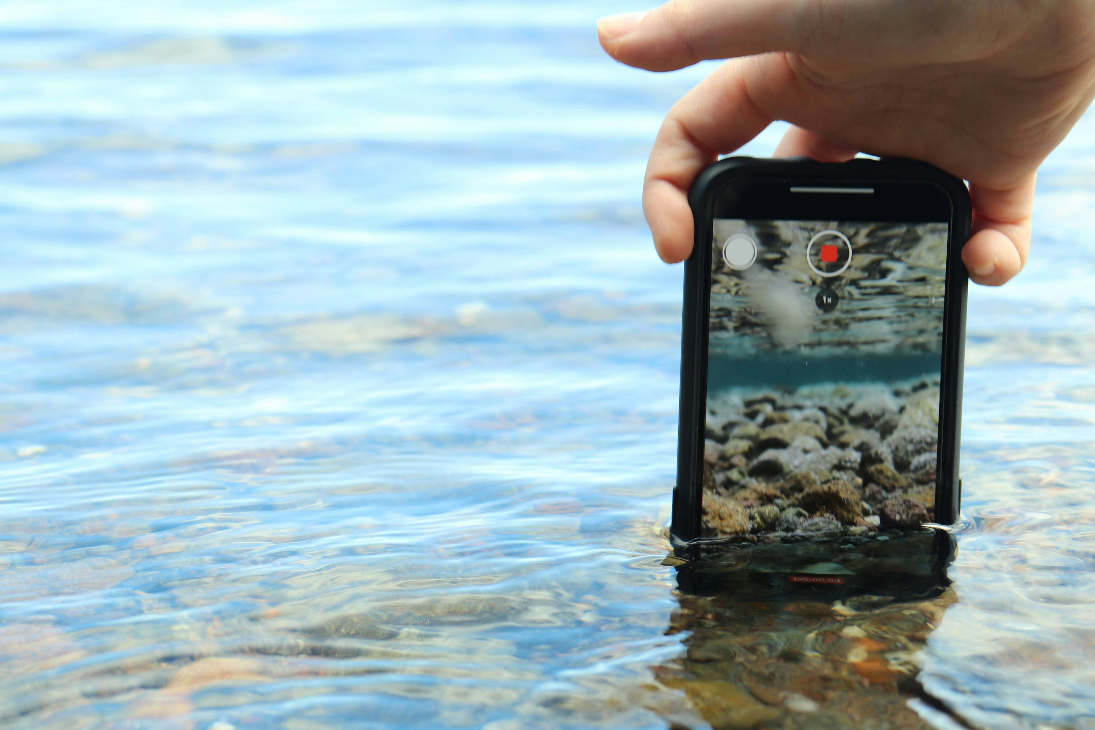 Hand hält ein Smartphone über Wasser mit sichtbaren Kieselsteinen darunter