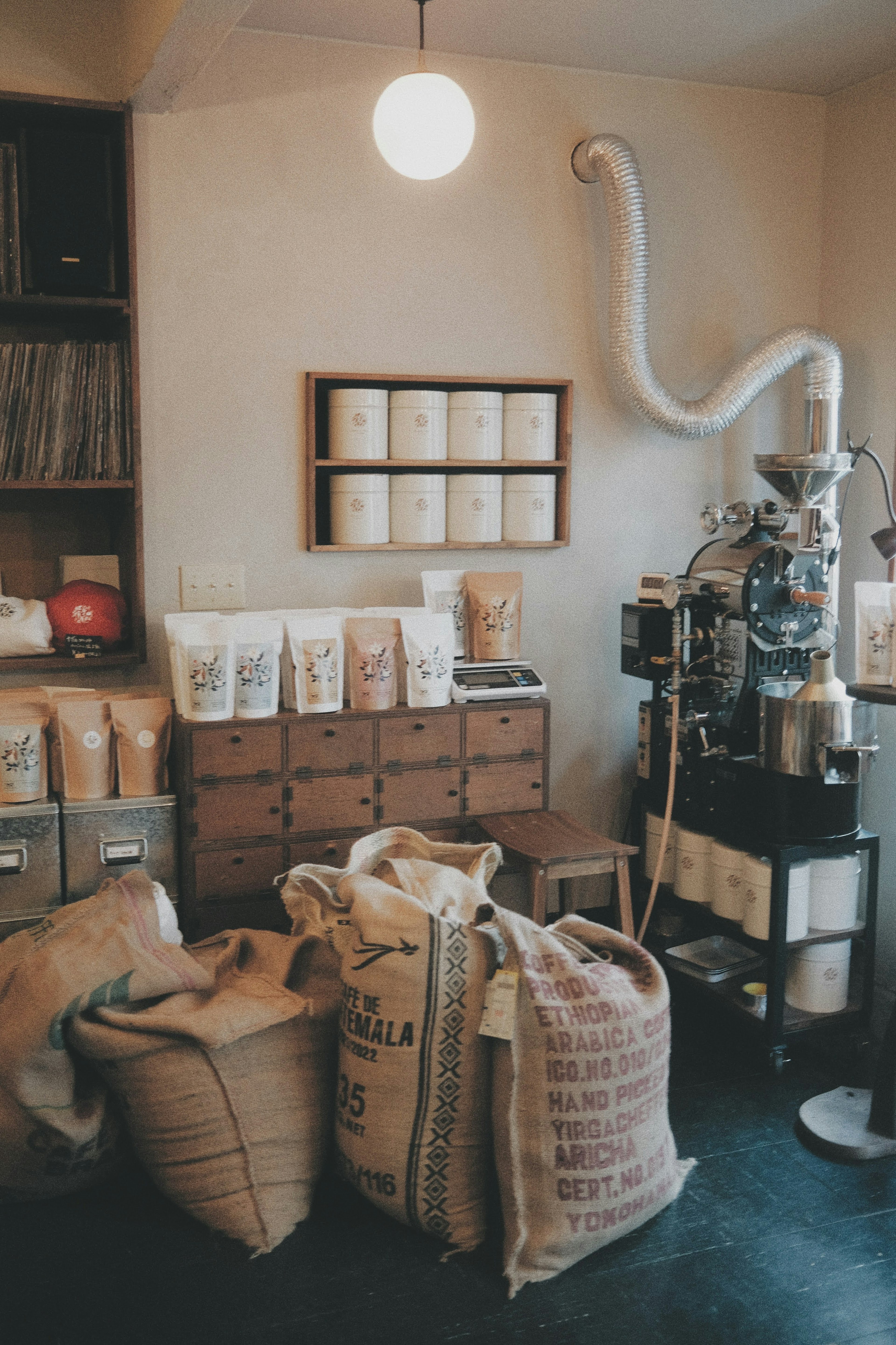 Interior of a coffee roasting shop featuring burlap sacks of coffee beans wooden drawers a roasting machine and simple lighting