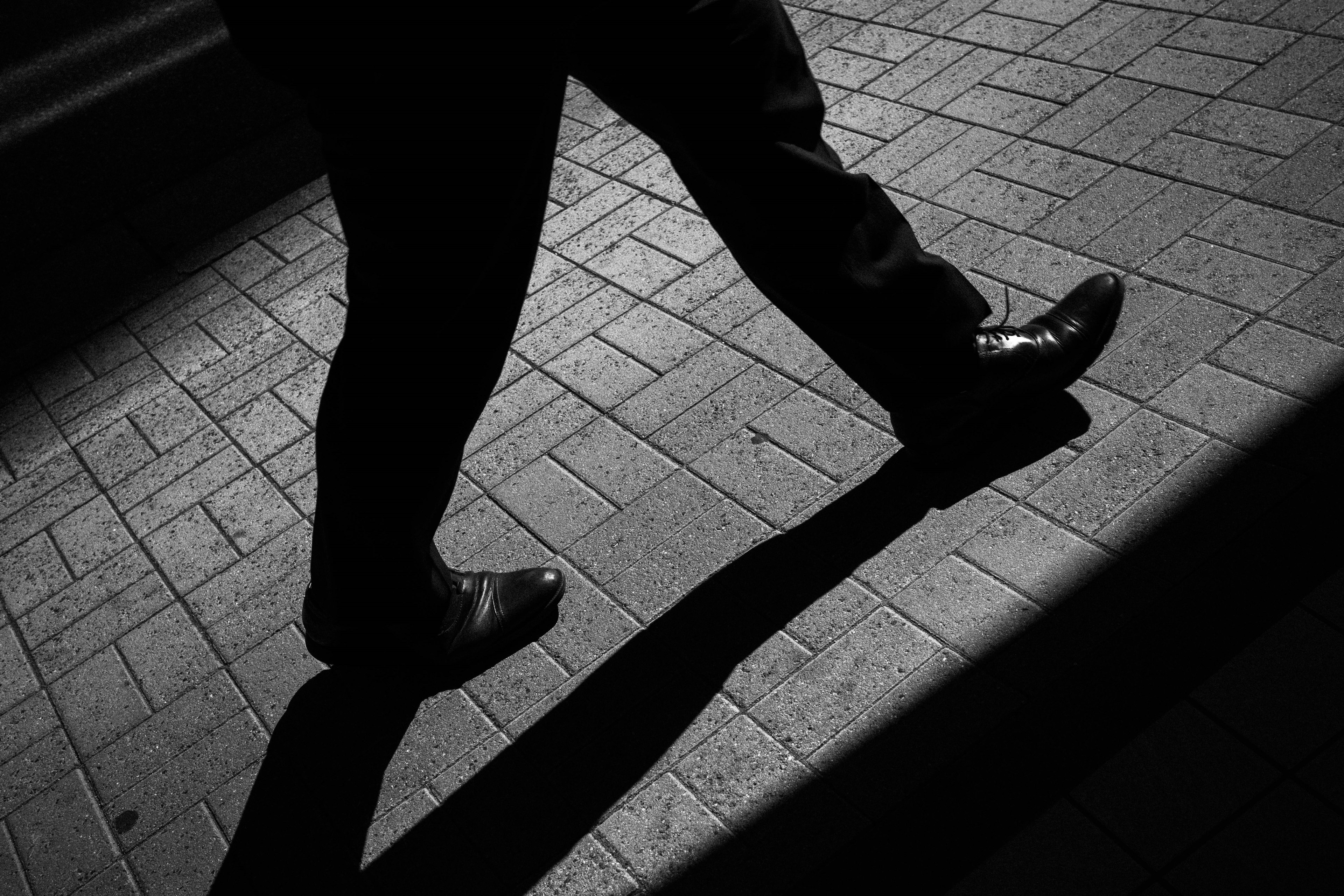 A man's foot in black shoes walking with a shadow on textured pavement