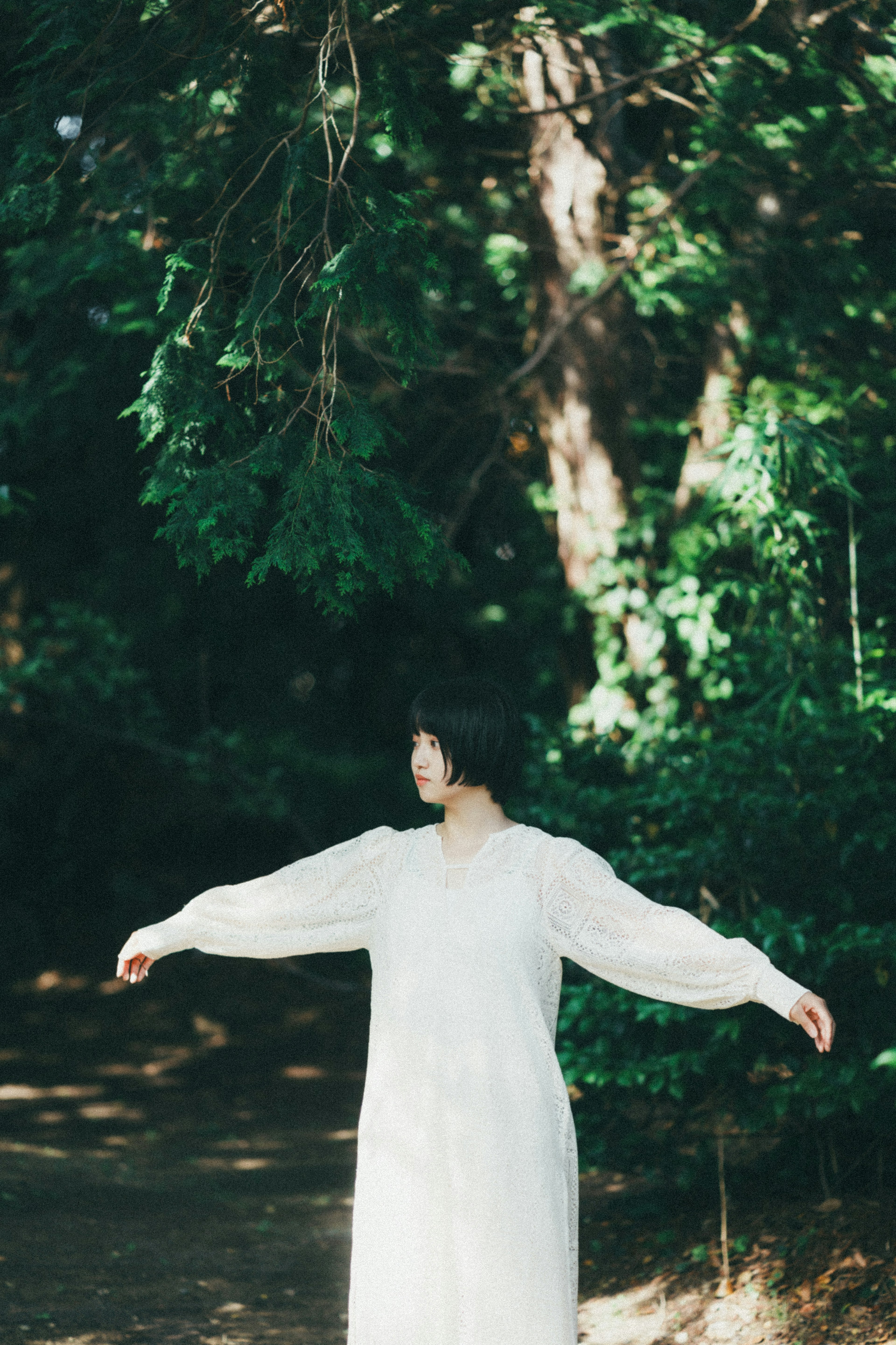 A woman in a white dress stands with arms outstretched in a forest