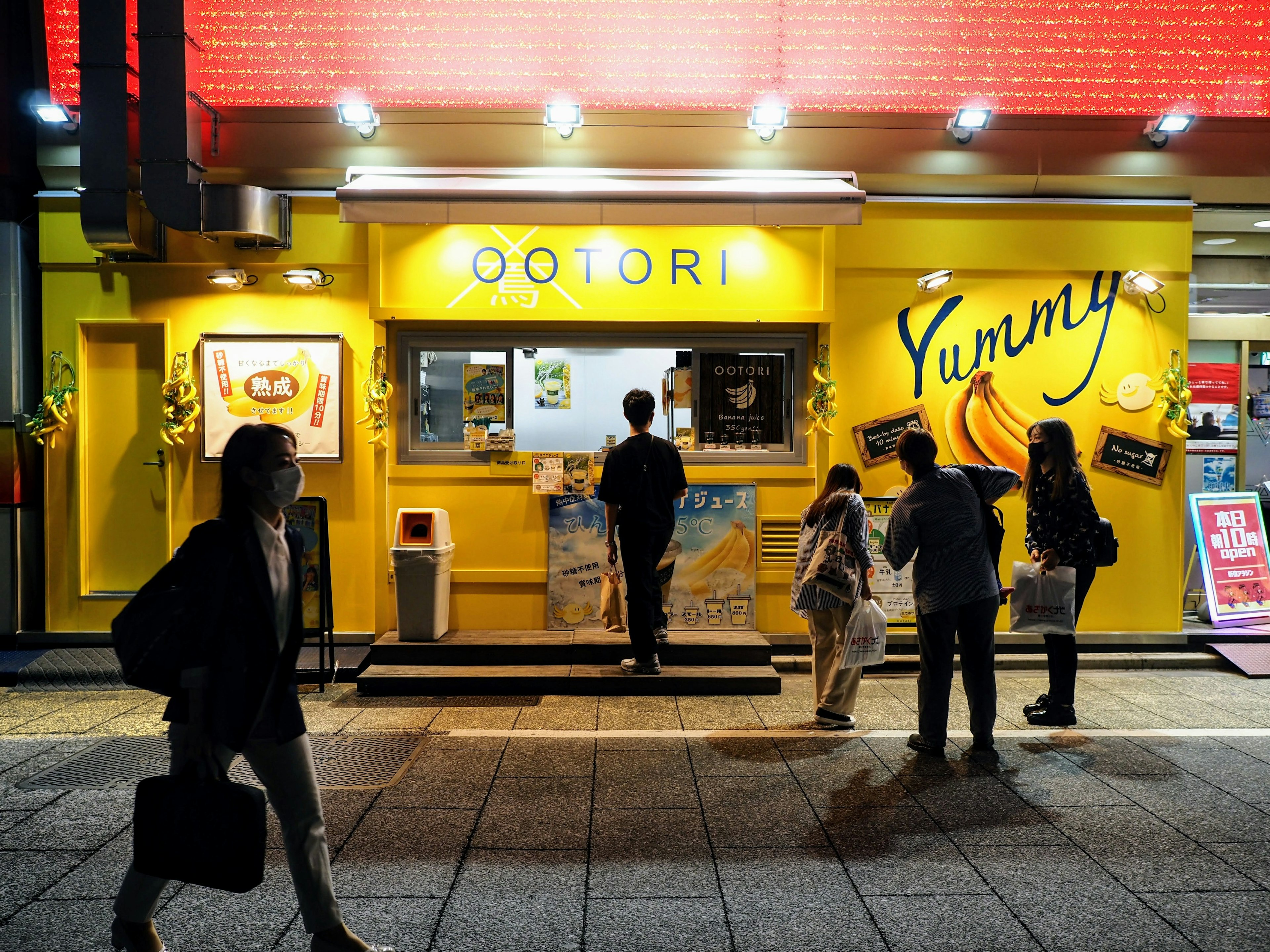 Des gens devant un magasin jaune avec un panneau 'Yummy' et un menu