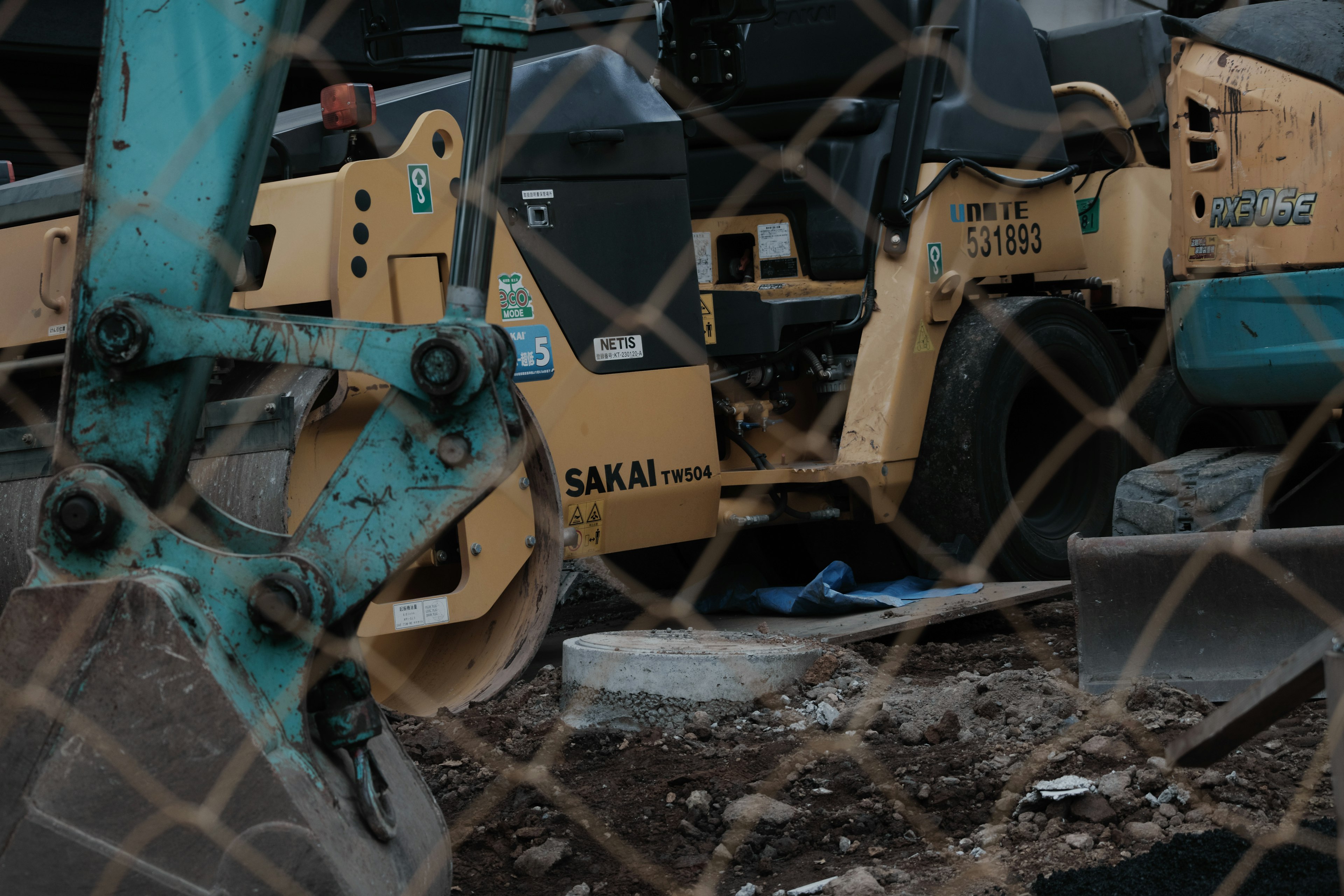 Part of a construction vehicle seen through a fence with surrounding dirt