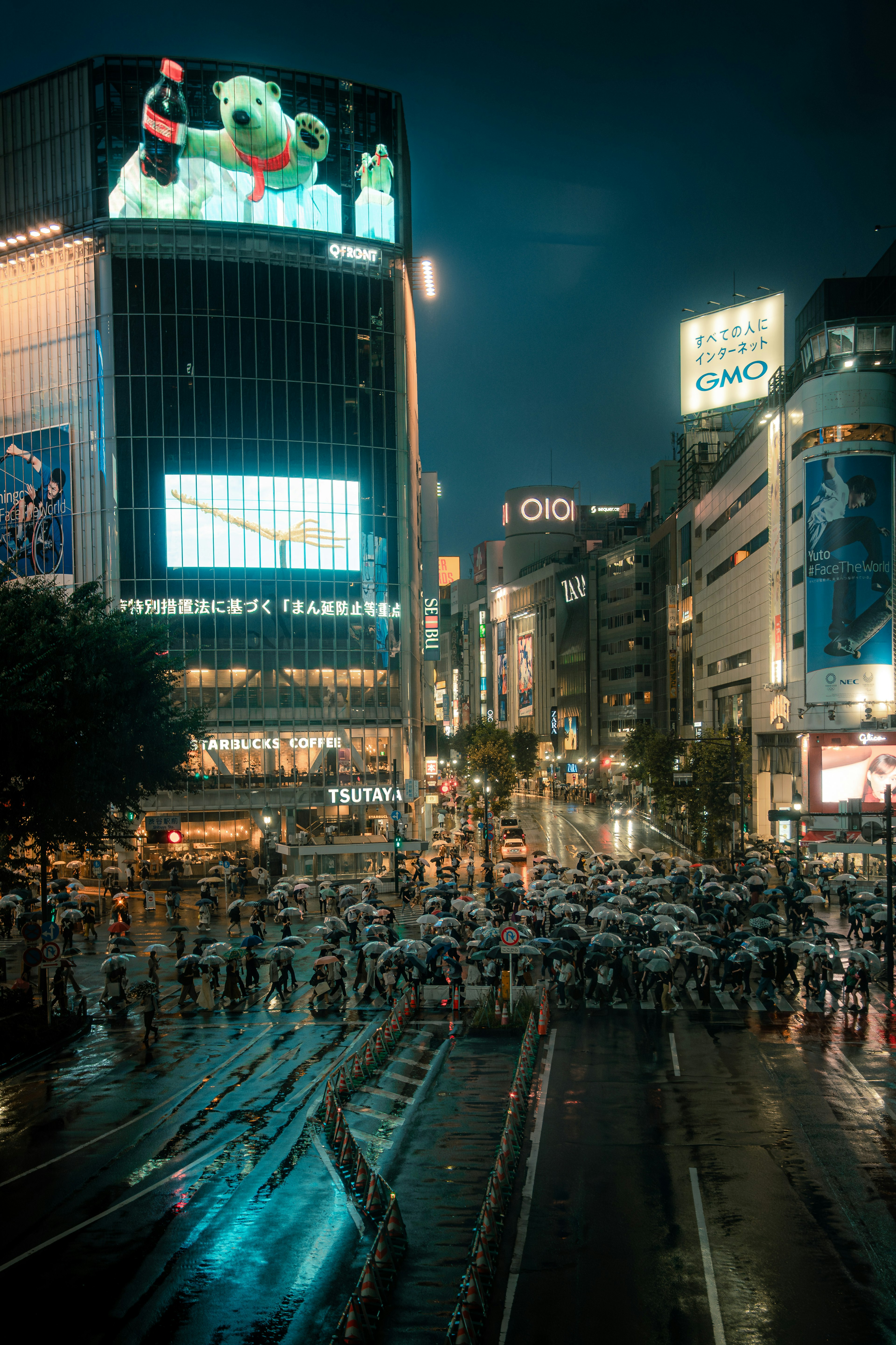 Persimpangan Shibuya yang sibuk di malam hari dengan layar besar menampilkan karakter dan orang-orang yang memegang payung