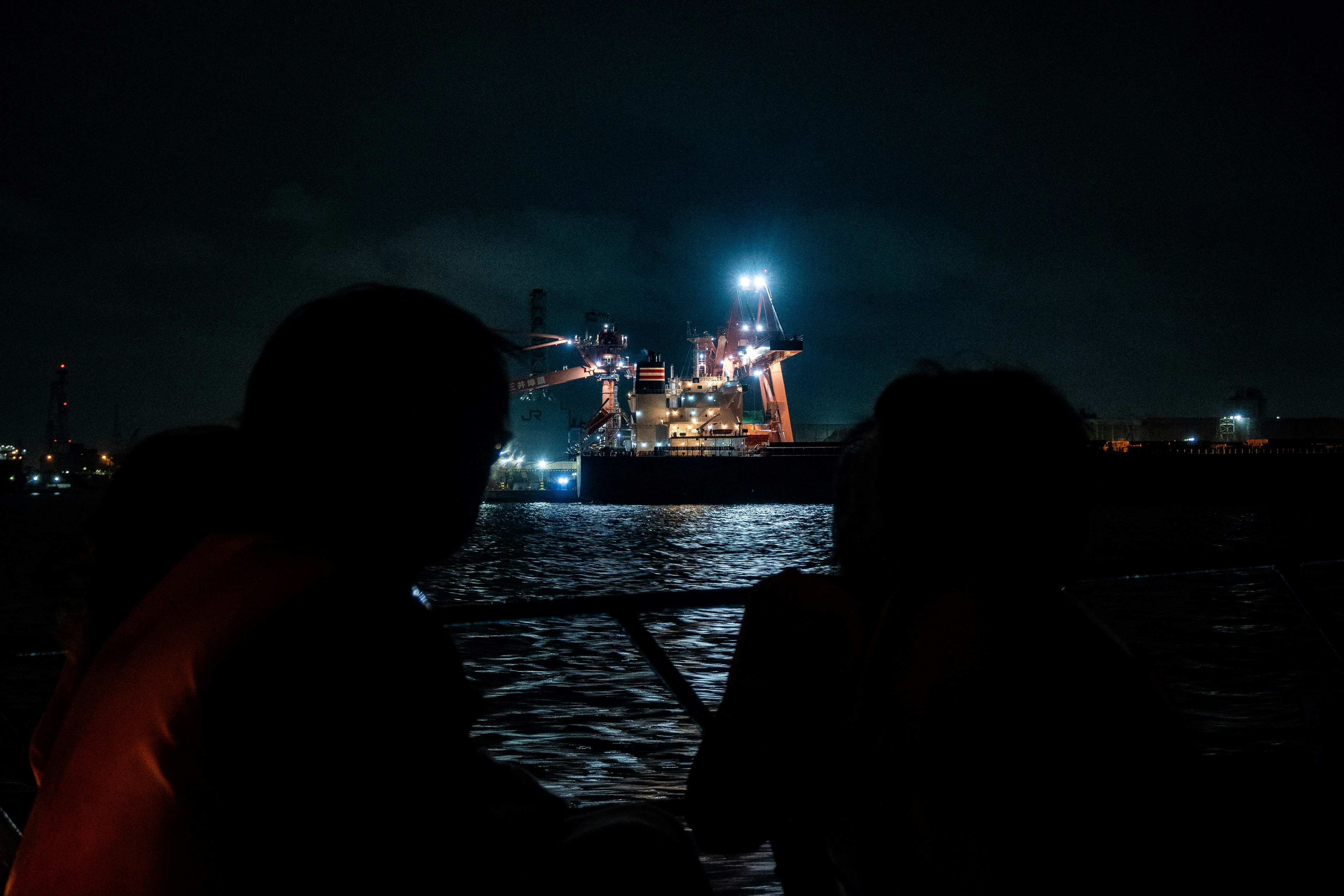 Siluetas de dos personas mirando un barco iluminado en el agua por la noche