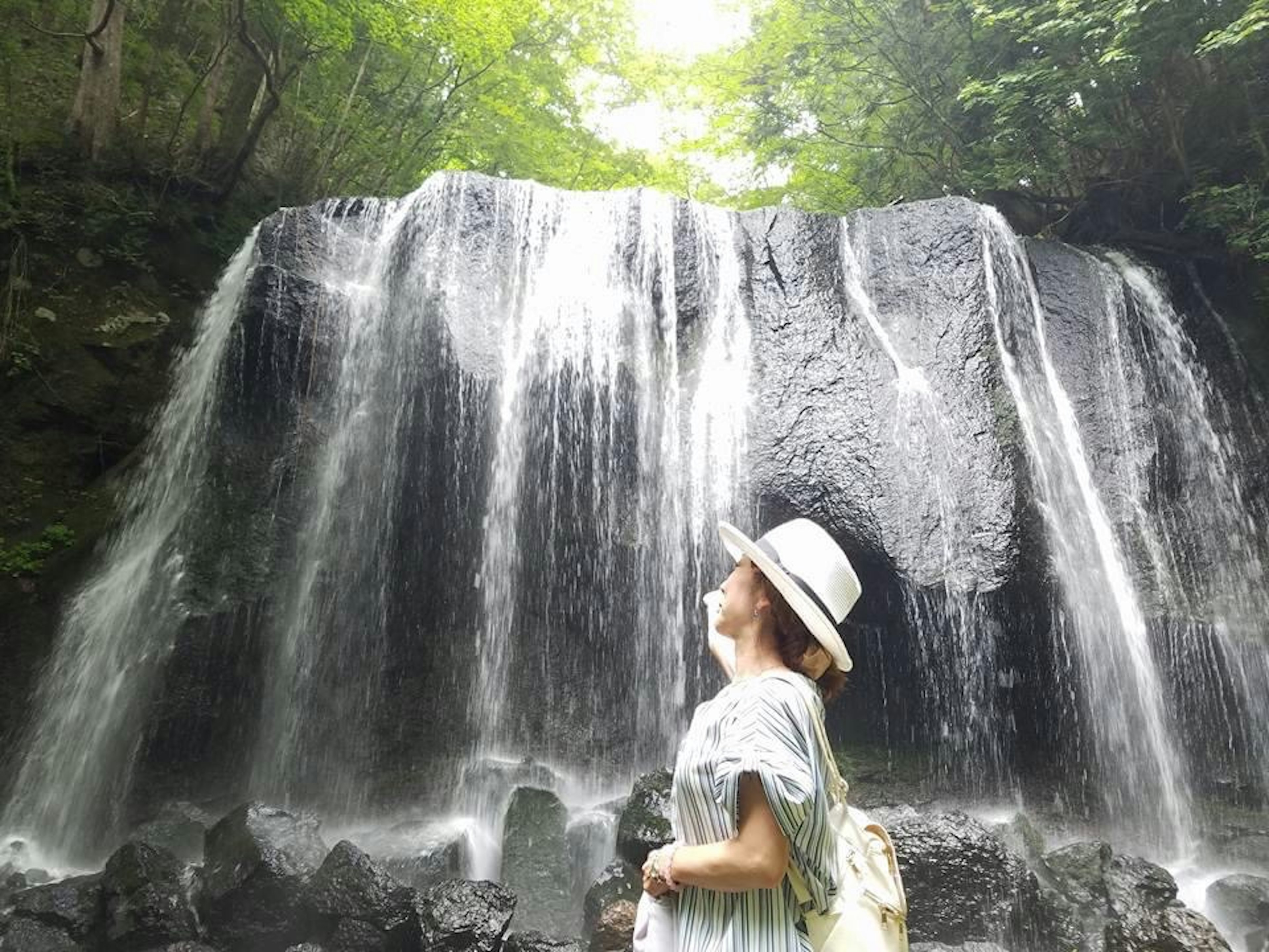 Seorang wanita mengenakan topi putih berdiri di depan air terjun menikmati keindahan alam