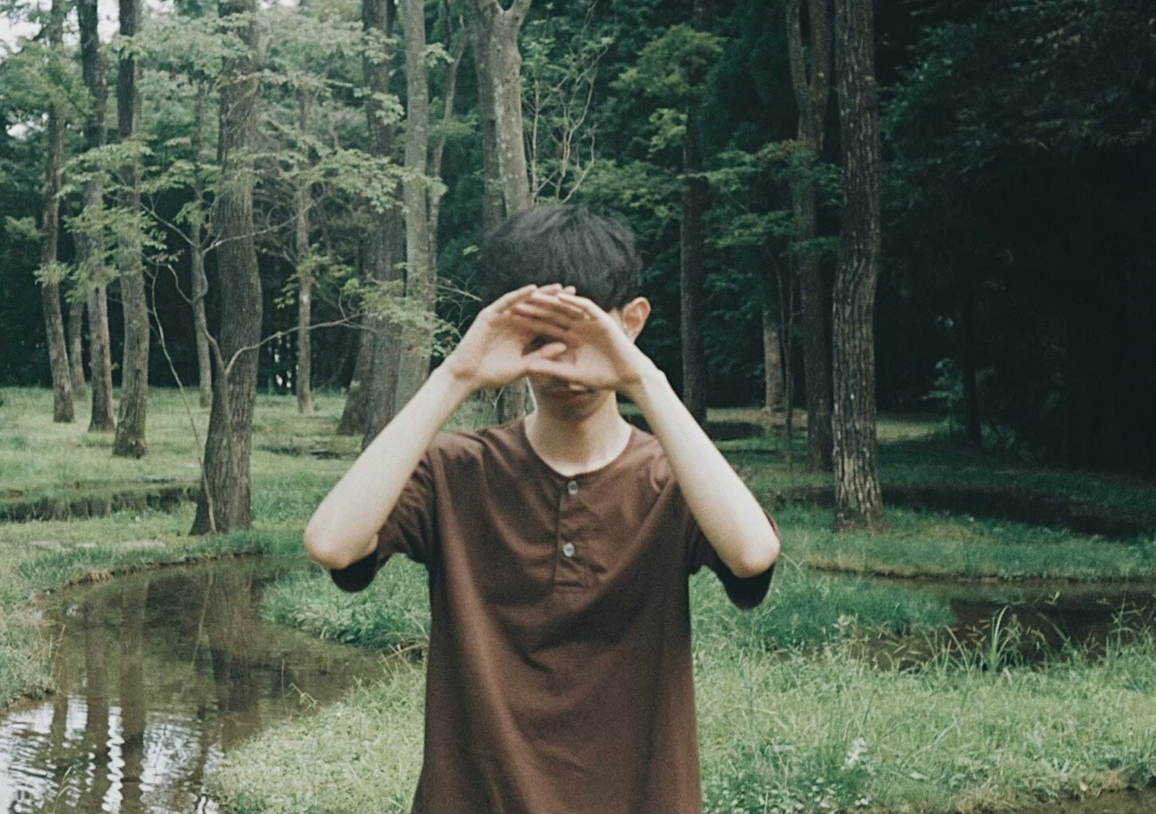 A young boy covering his eyes with his hands in a green forest