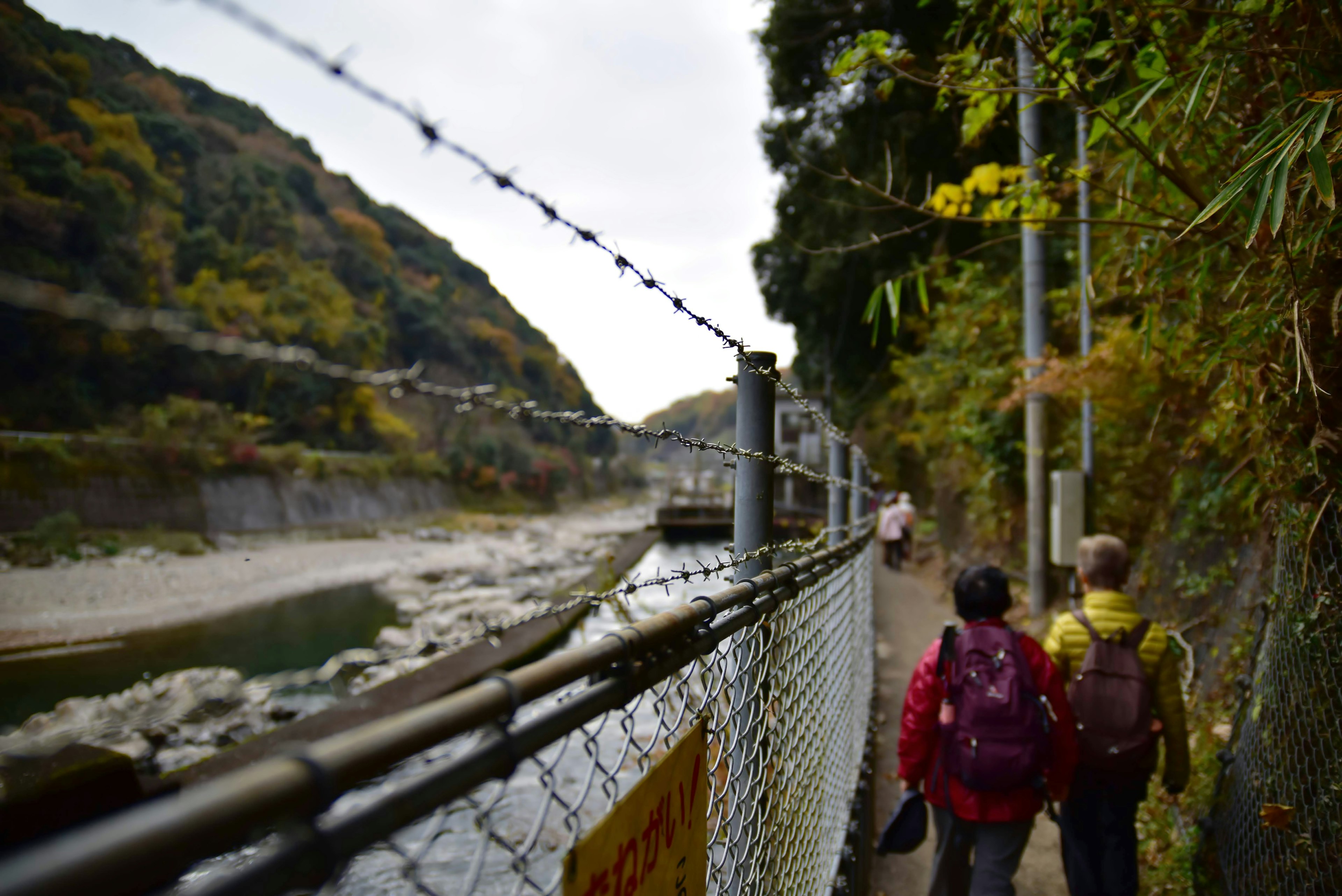 People walking along a path near a river with a barbed wire fence and lush greenery