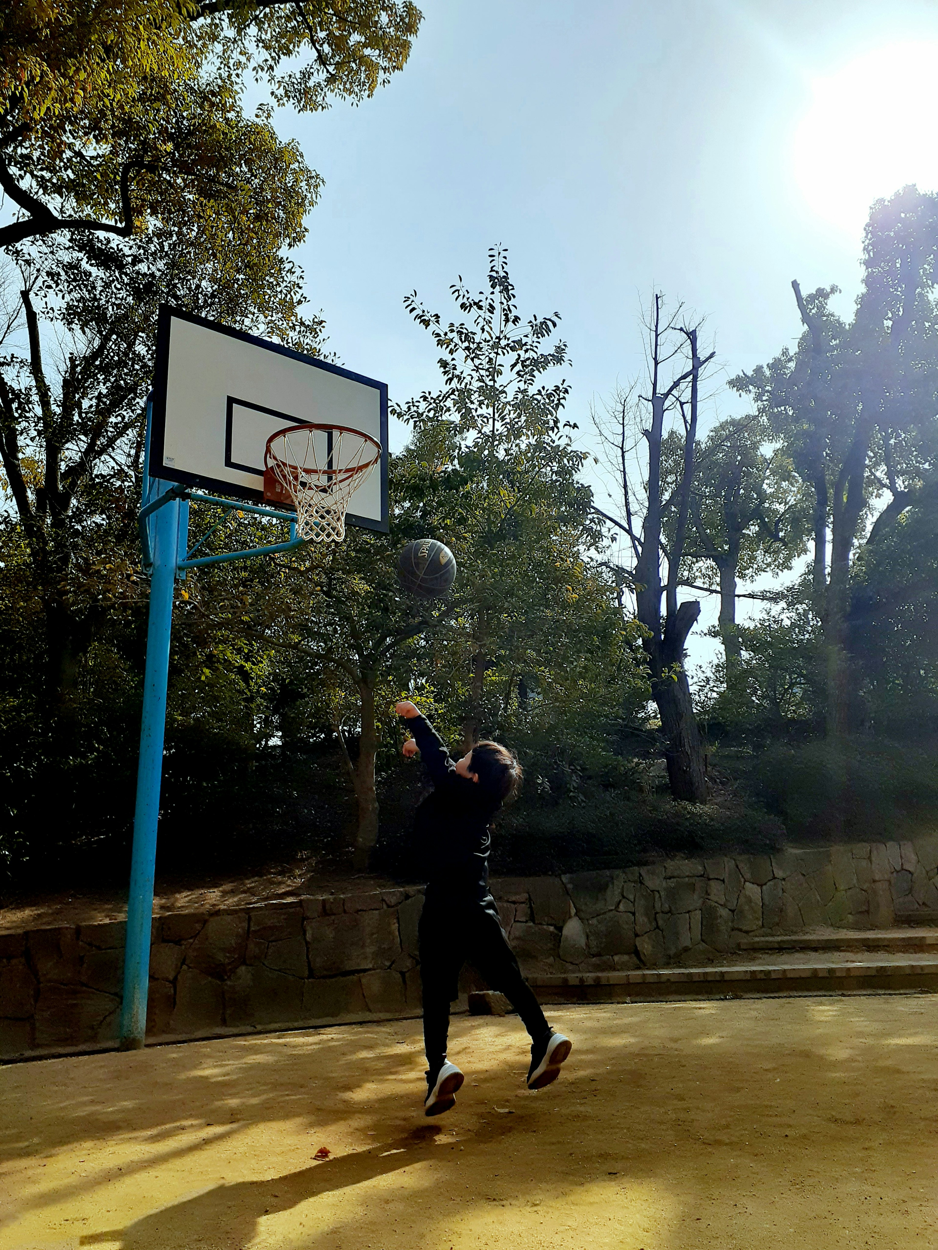 Seorang anak laki-laki melempar bola basket dengan latar belakang langit biru dan pohon hijau