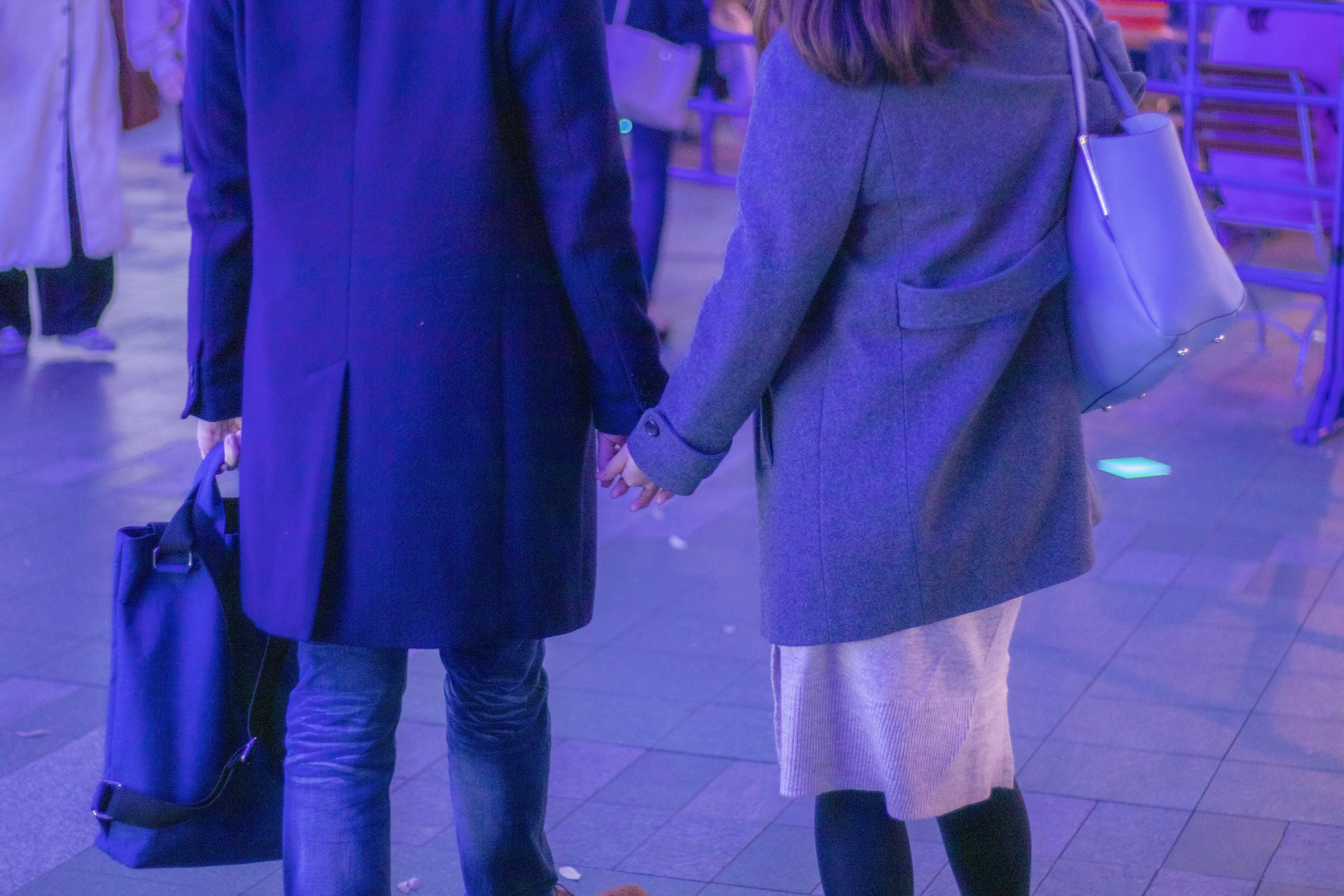 Couple holding hands walking in a dimly lit area wearing blue and gray coats