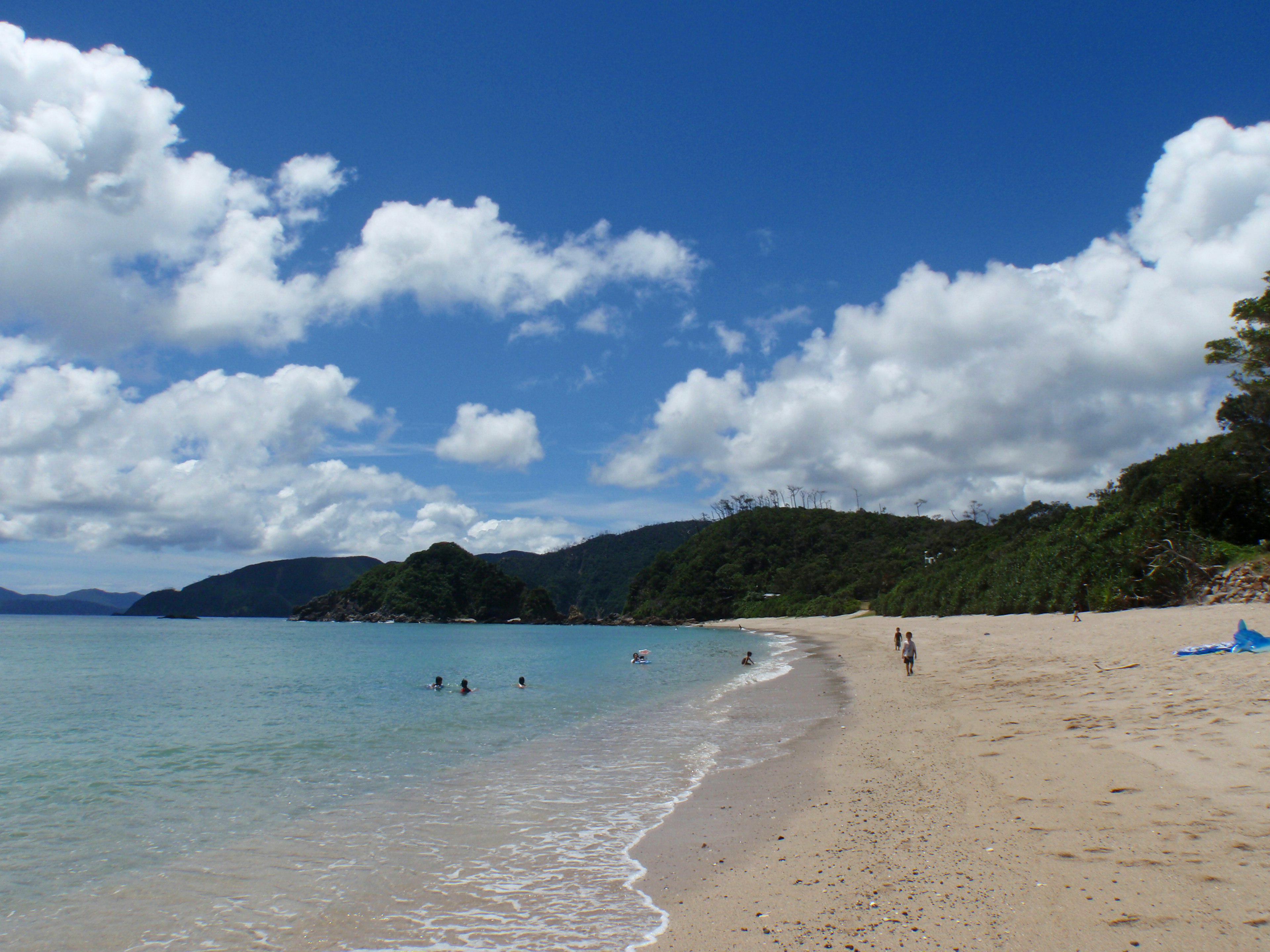 Escena de playa con personas disfrutando del agua bajo un cielo azul y nubes blancas