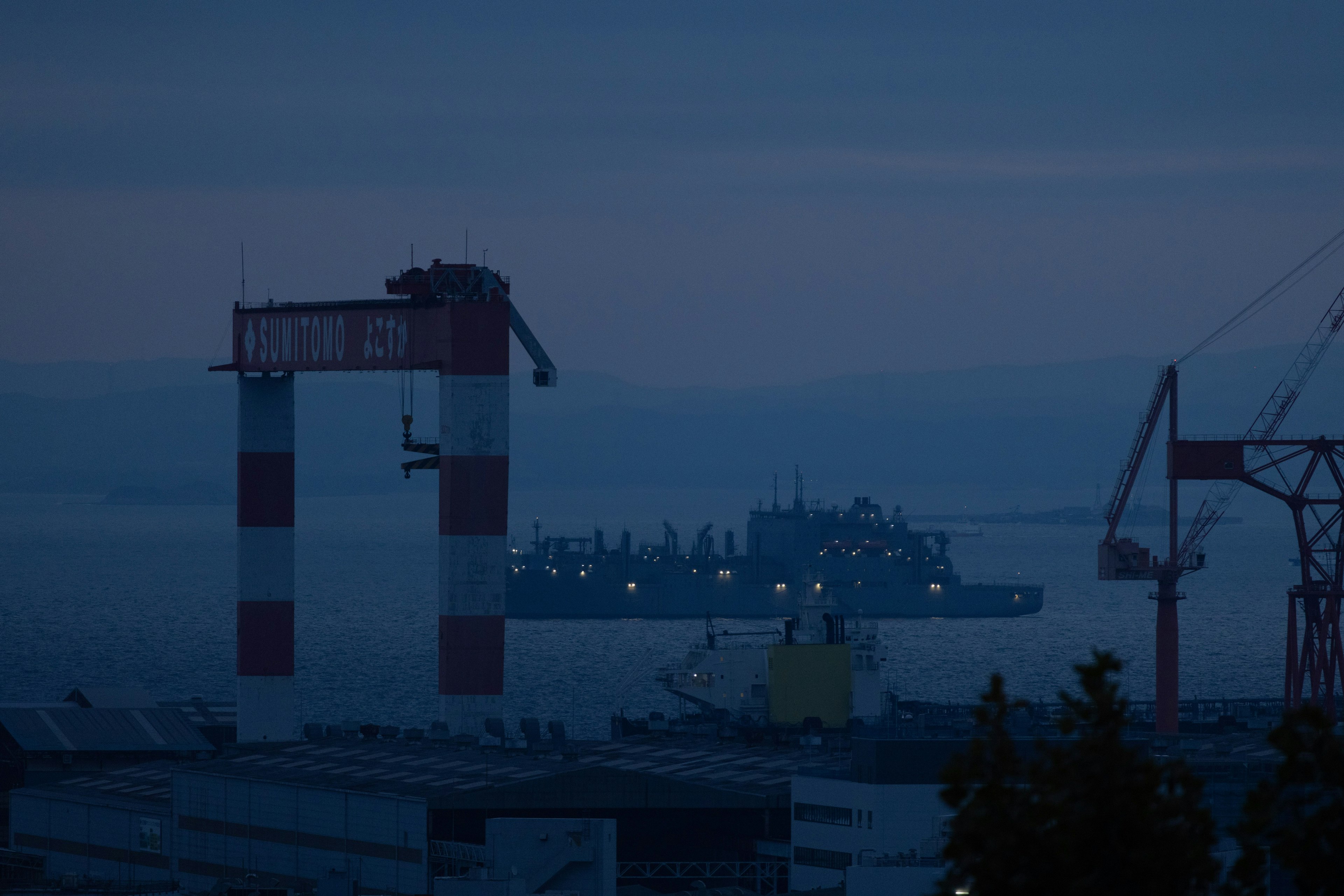 Silhouette von rot-weißen Kränen und einem Schiff in einem schwach beleuchteten Hafen bei Nacht