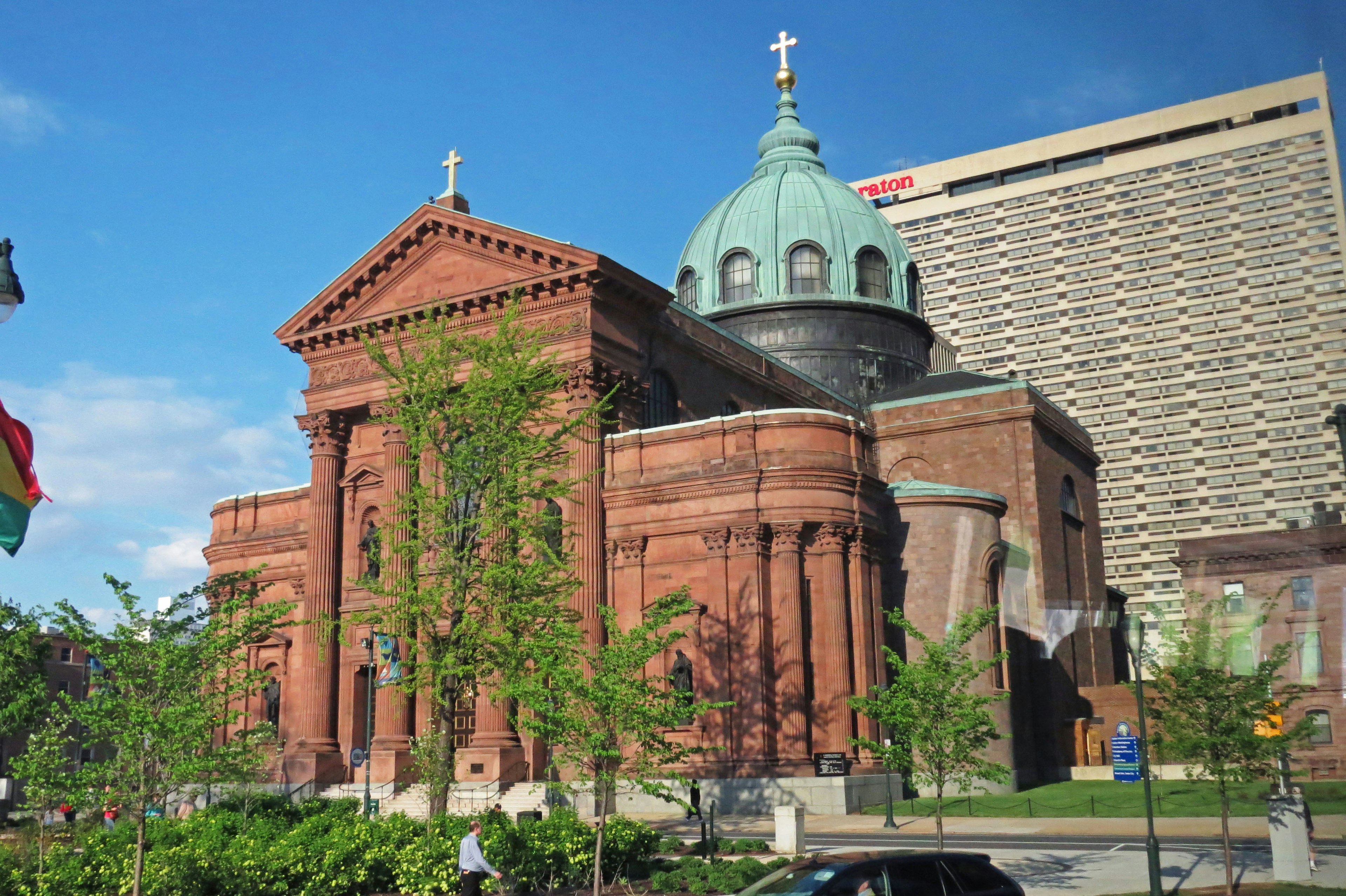 Red brick church featuring a green dome with a delicate cross on top surrounded by green trees and modern buildings