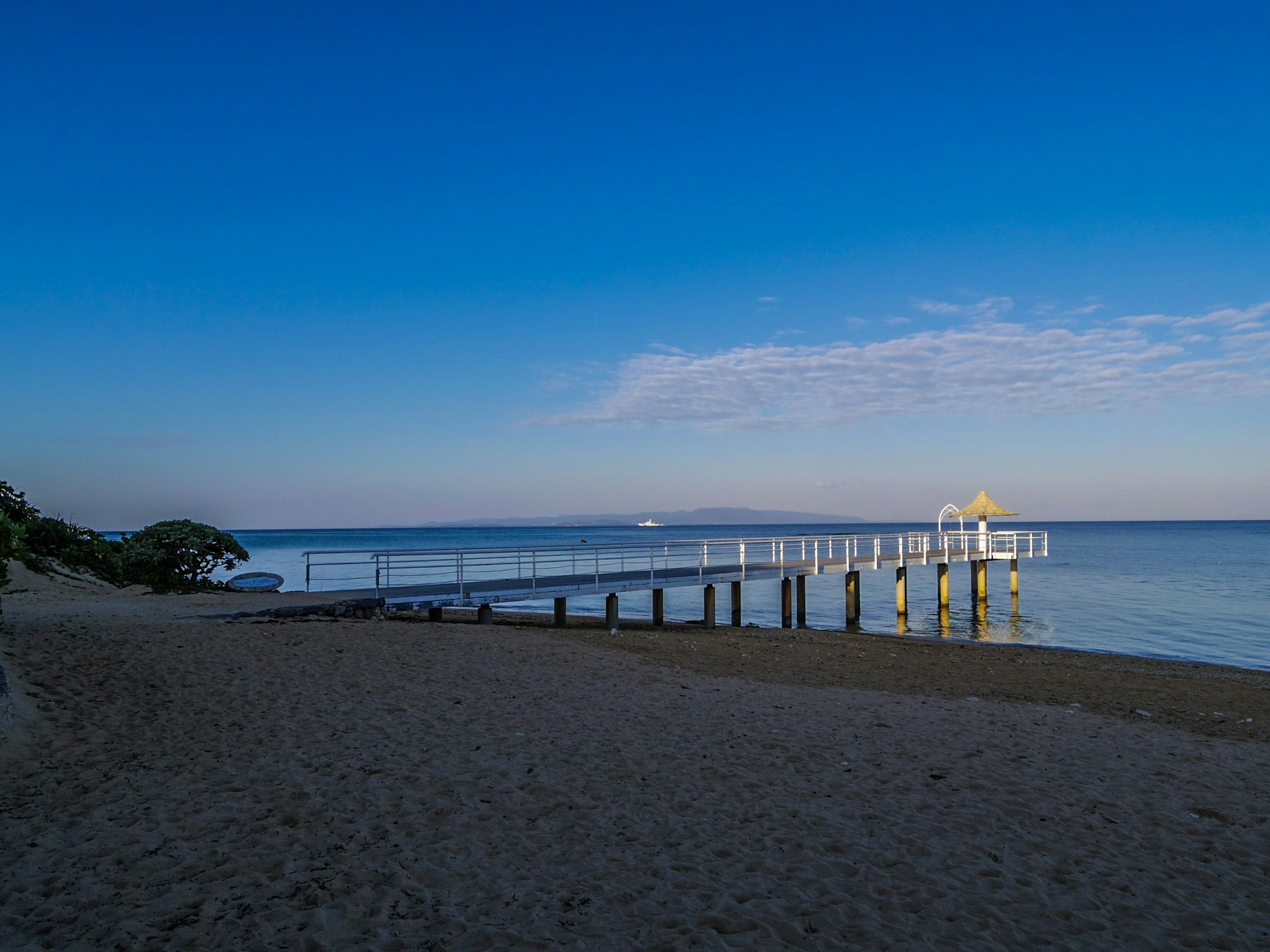 ภาพชายหาดที่มีท่าเทียบเรือยื่นออกไปสู่ทะเลสีน้ำเงินใสใต้ท้องฟ้าที่ชัดเจน