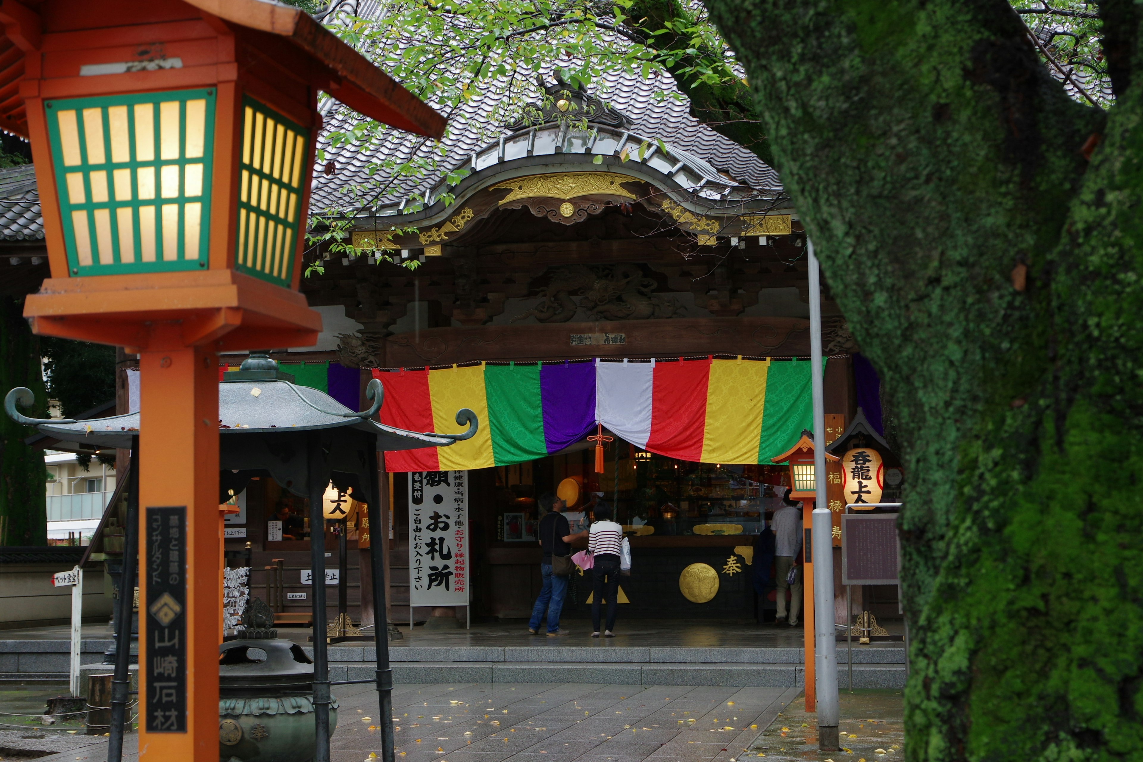 Entrance of a shrine adorned with colorful banners, featuring an orange lantern and lush green trees