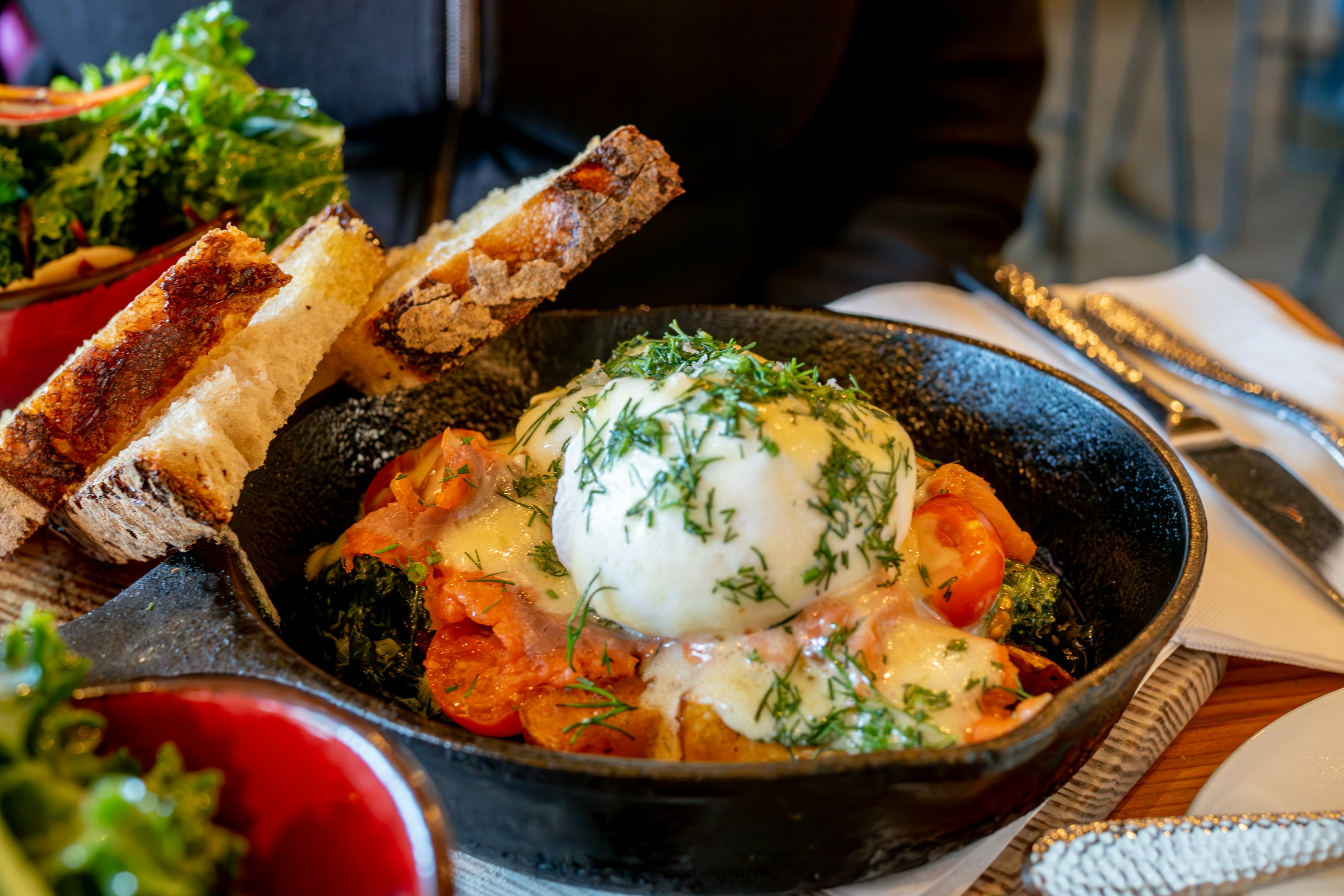 Plato de huevos servido en una sartén, cubierto con hierbas frescas, acompañado de pan tostado y ensalada