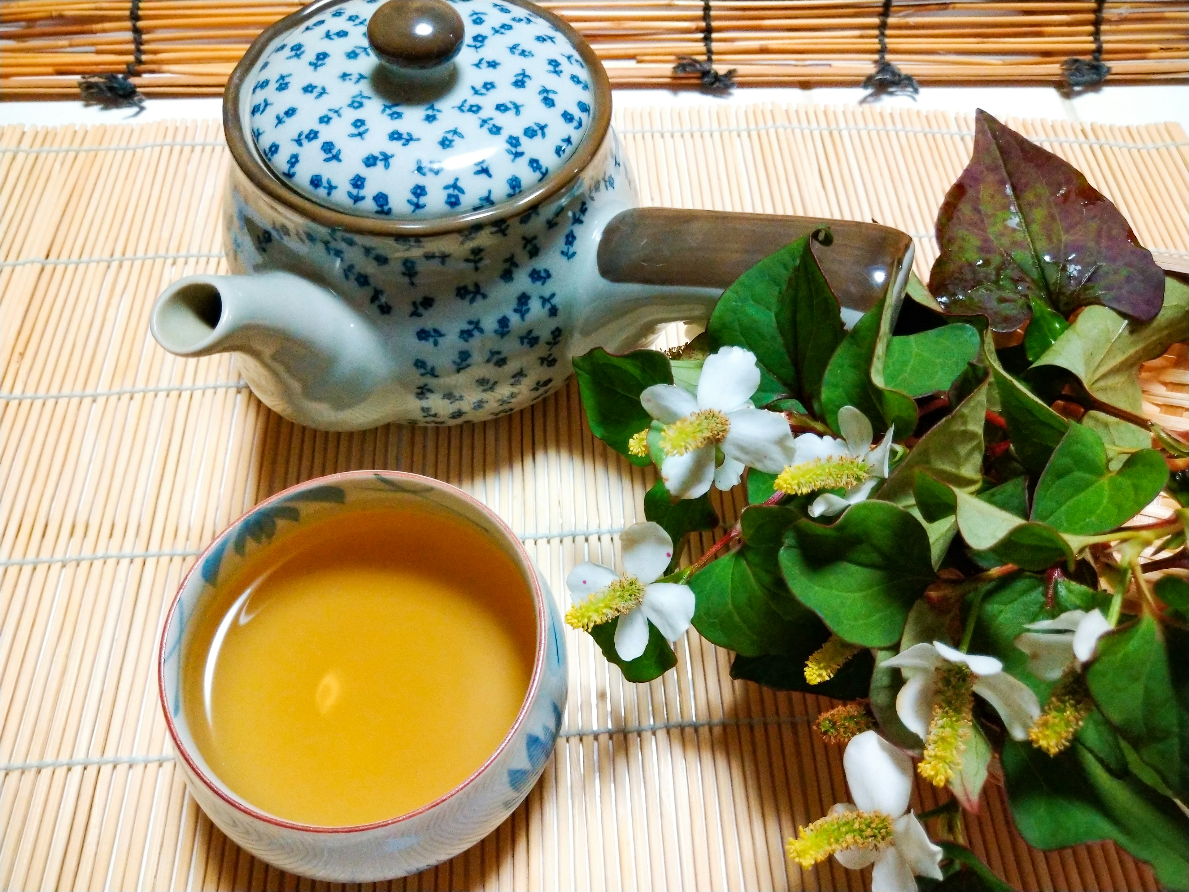 Blue patterned teapot next to a cup of yellow tea and green leaves on a bamboo mat