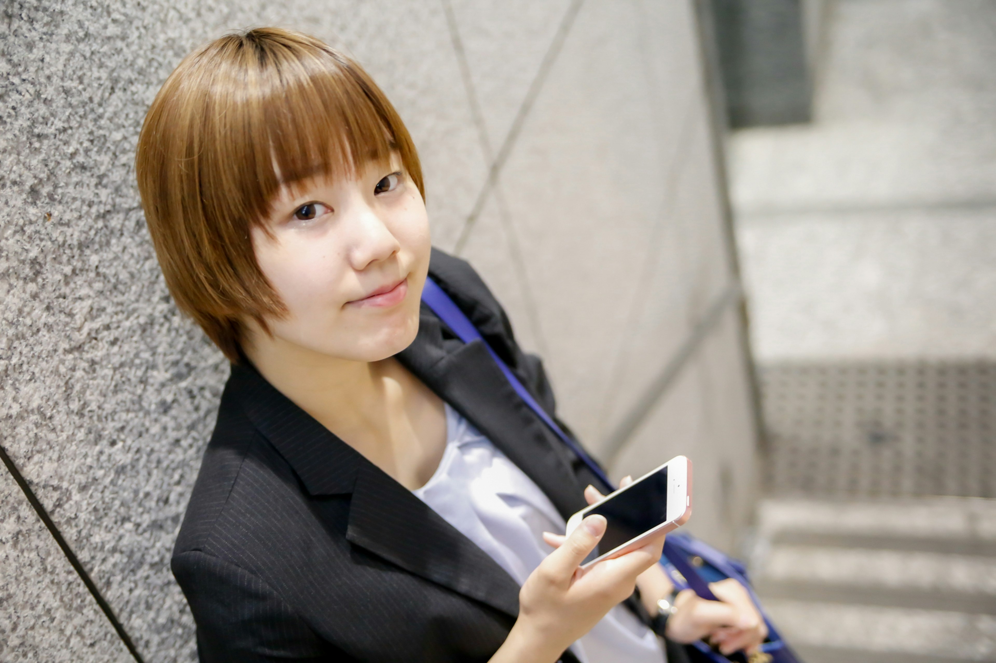 A short-haired woman leaning against a staircase holding a smartphone and smiling