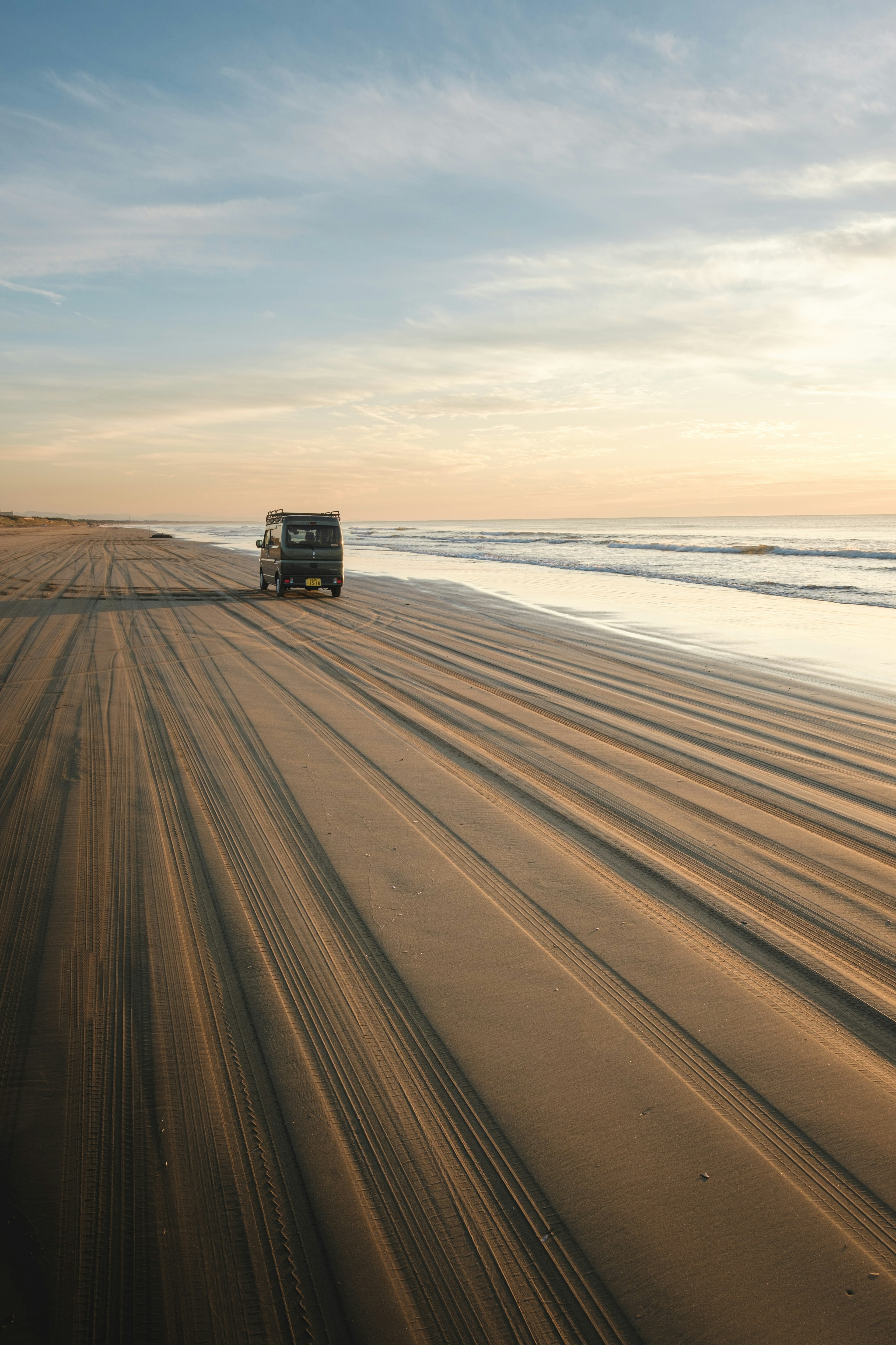 Ein Fahrzeug fährt über einen Sandstrand mit Reifenspuren und einem Sonnenuntergangshimmel