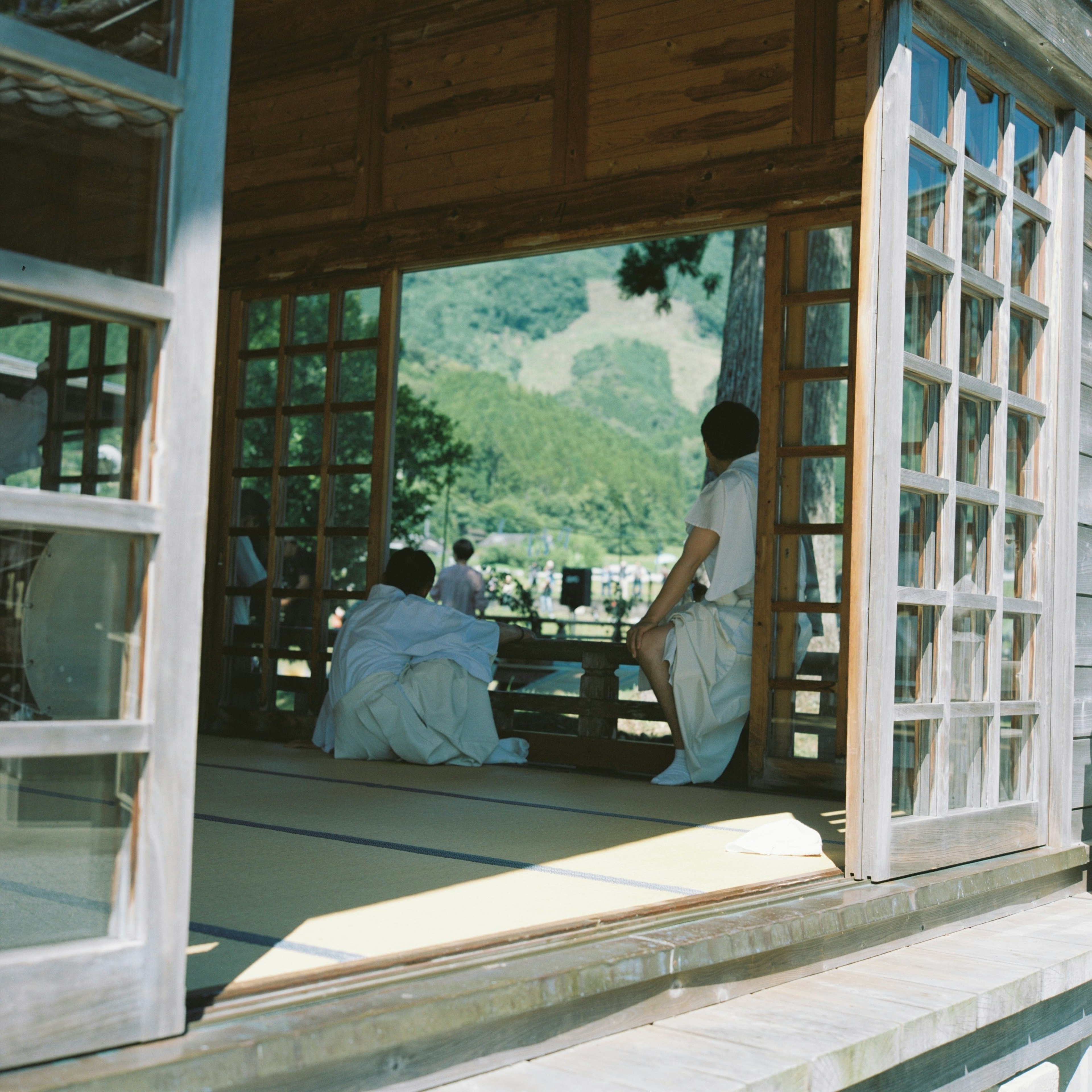 木造の日本の建物の内部で、座っている人物が見える緑豊かな風景