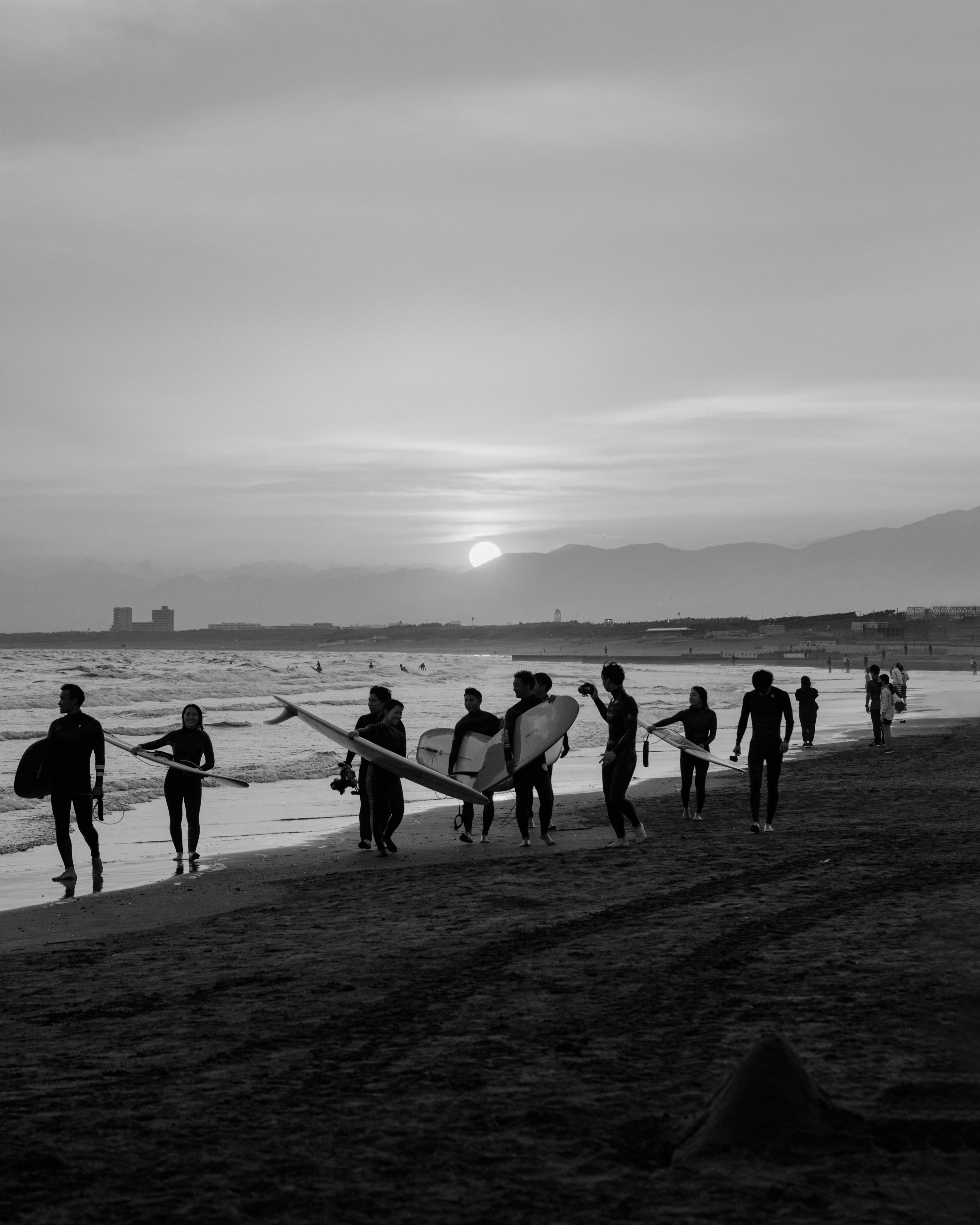 Foto in bianco e nero di surfisti che camminano lungo la spiaggia al tramonto