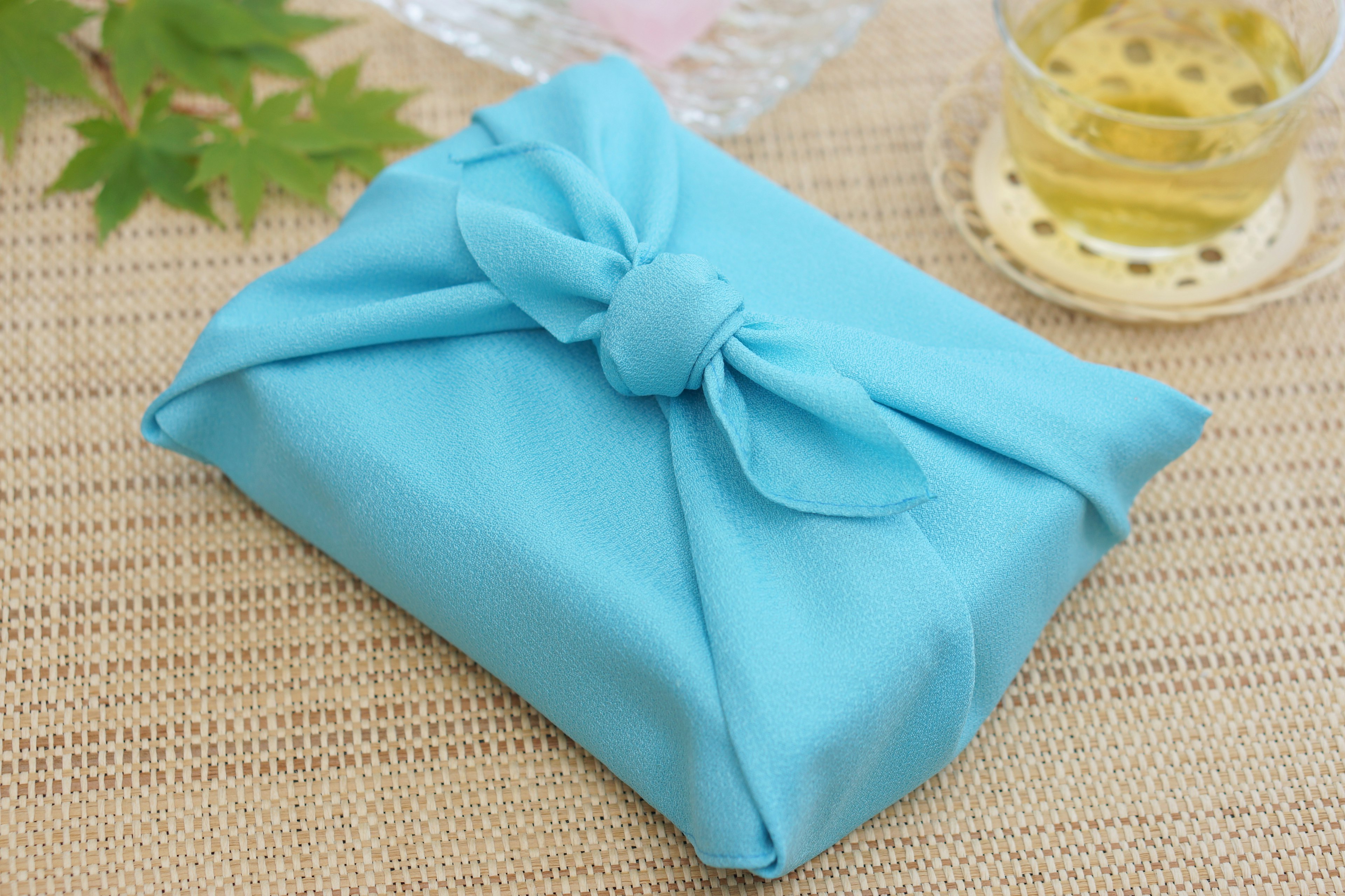 A gift box wrapped in blue fabric placed next to green leaves and tea