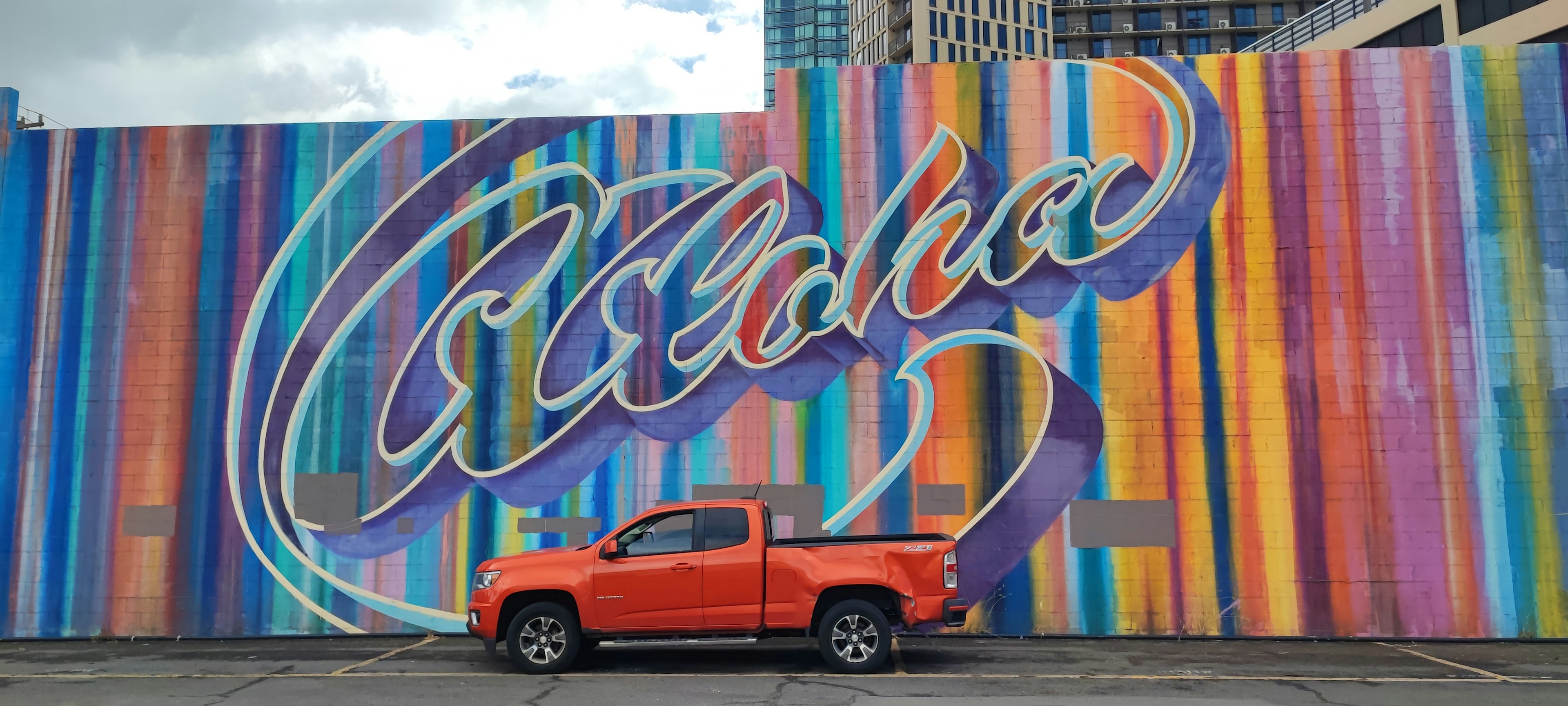 Una camioneta naranja estacionada frente a un mural colorido con la palabra Aloha