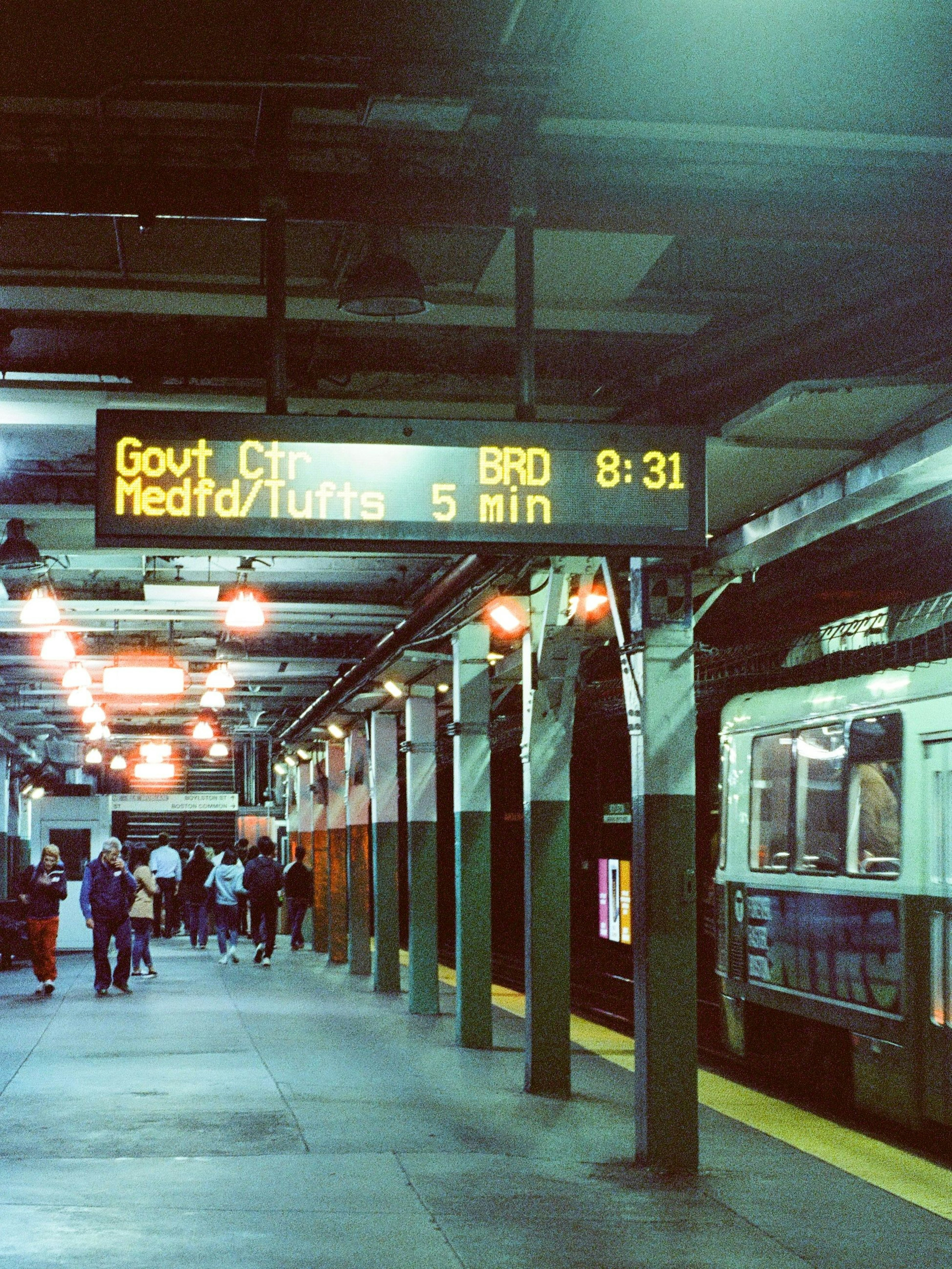 Personas esperando en una plataforma de metro con un panel digital que muestra información del tren