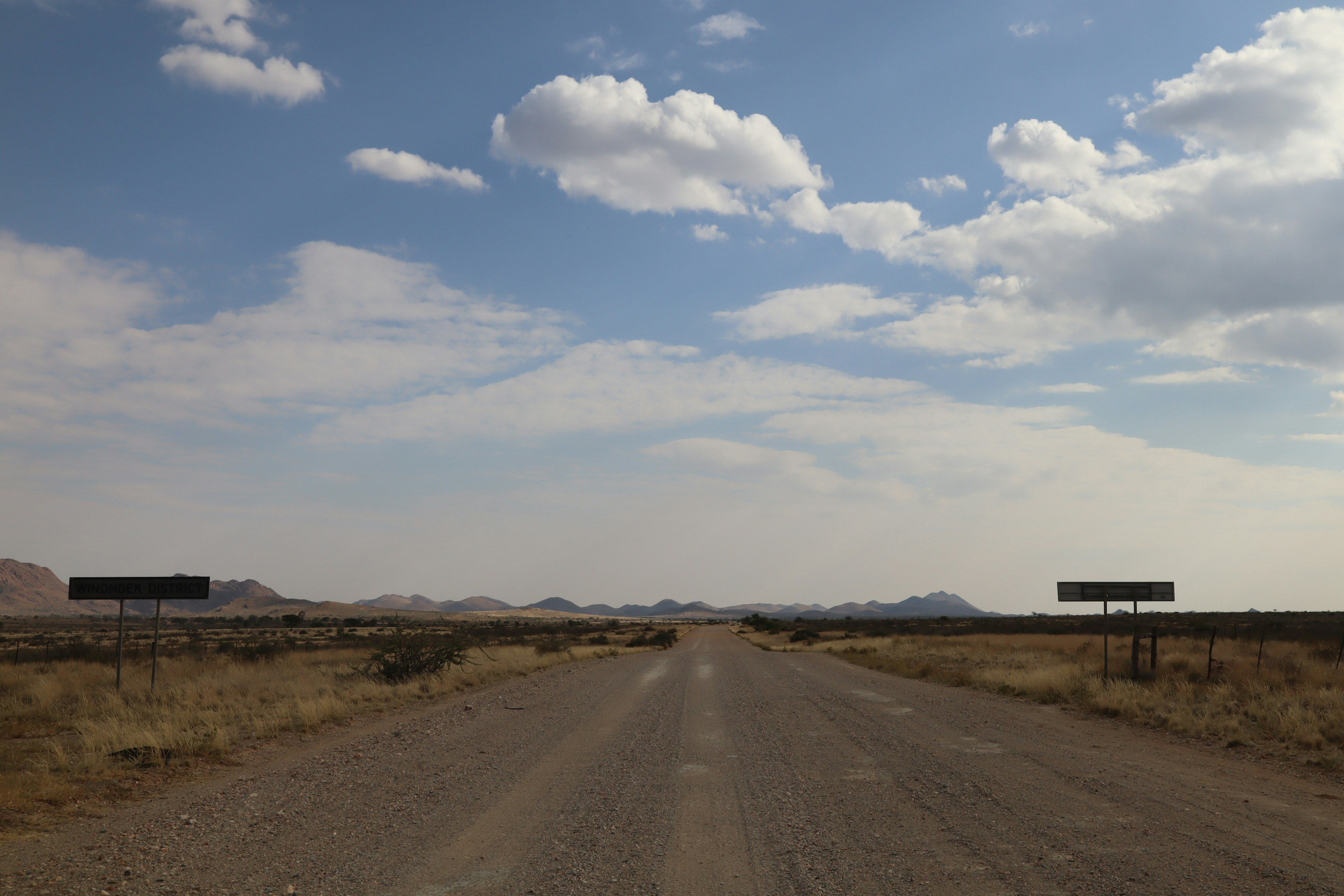 Wide gravel road under a blue sky with distant mountains and clouds