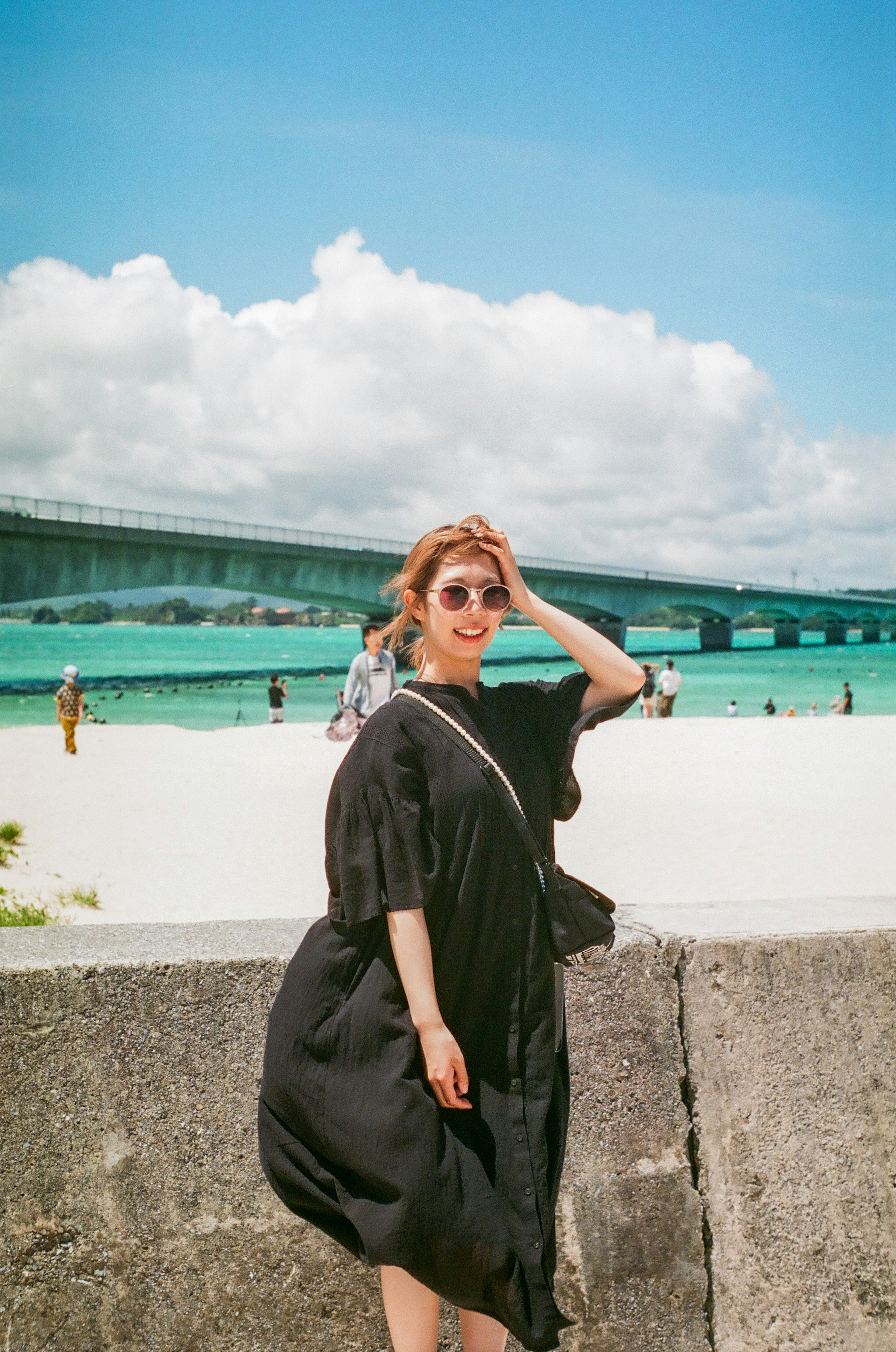 Femme en robe noire posant sur la plage avec un pont en arrière-plan