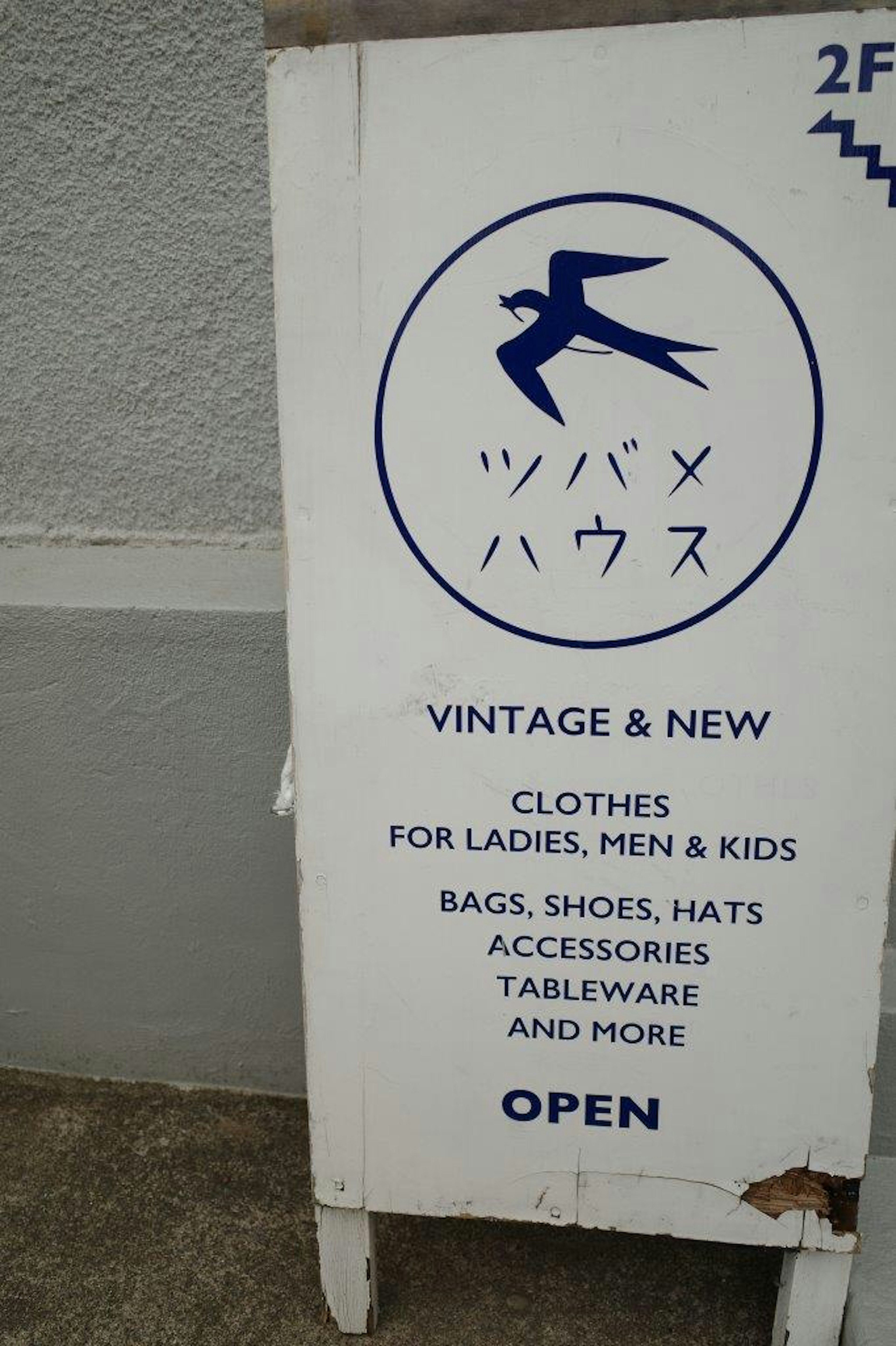 Panneau d'un magasin de vêtements vintage et neufs proposant des vêtements pour dames, hommes et enfants ainsi que des sacs, chaussures, chapeaux, accessoires et vaisselle