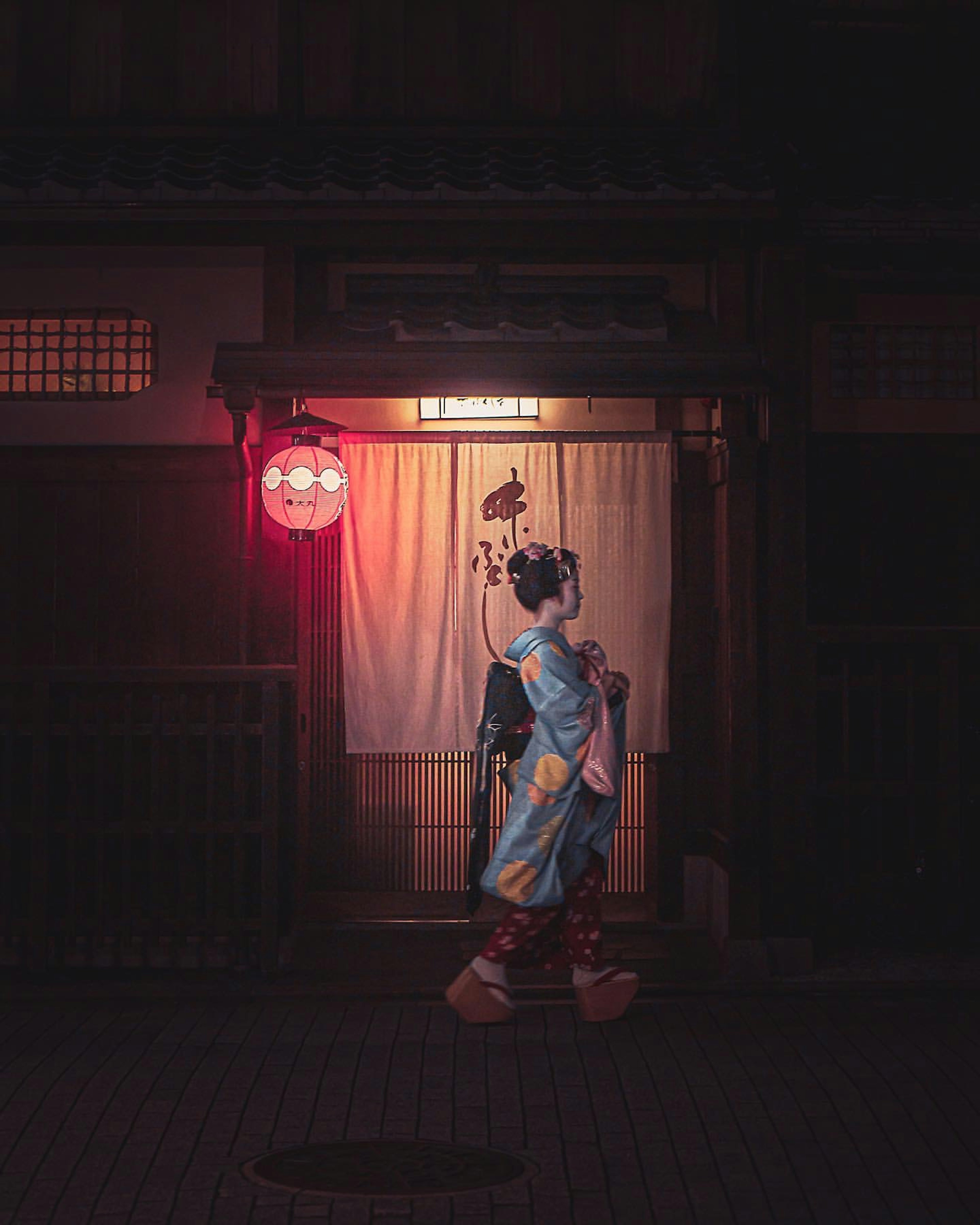 Una mujer en kimono caminando bajo una linterna roja en una escena nocturna