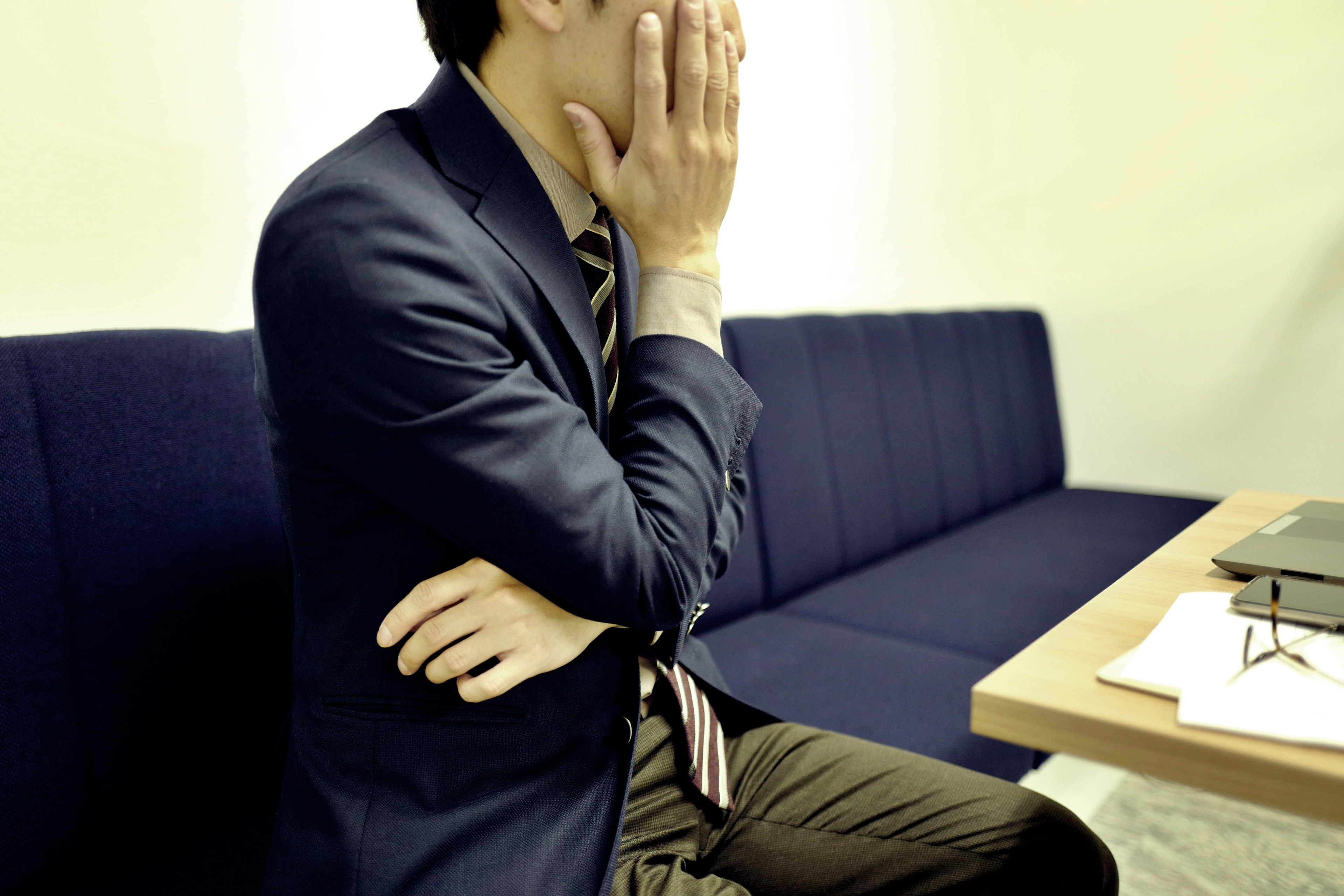 A man in a business suit sitting on a couch covering his face with his hand looking pensive