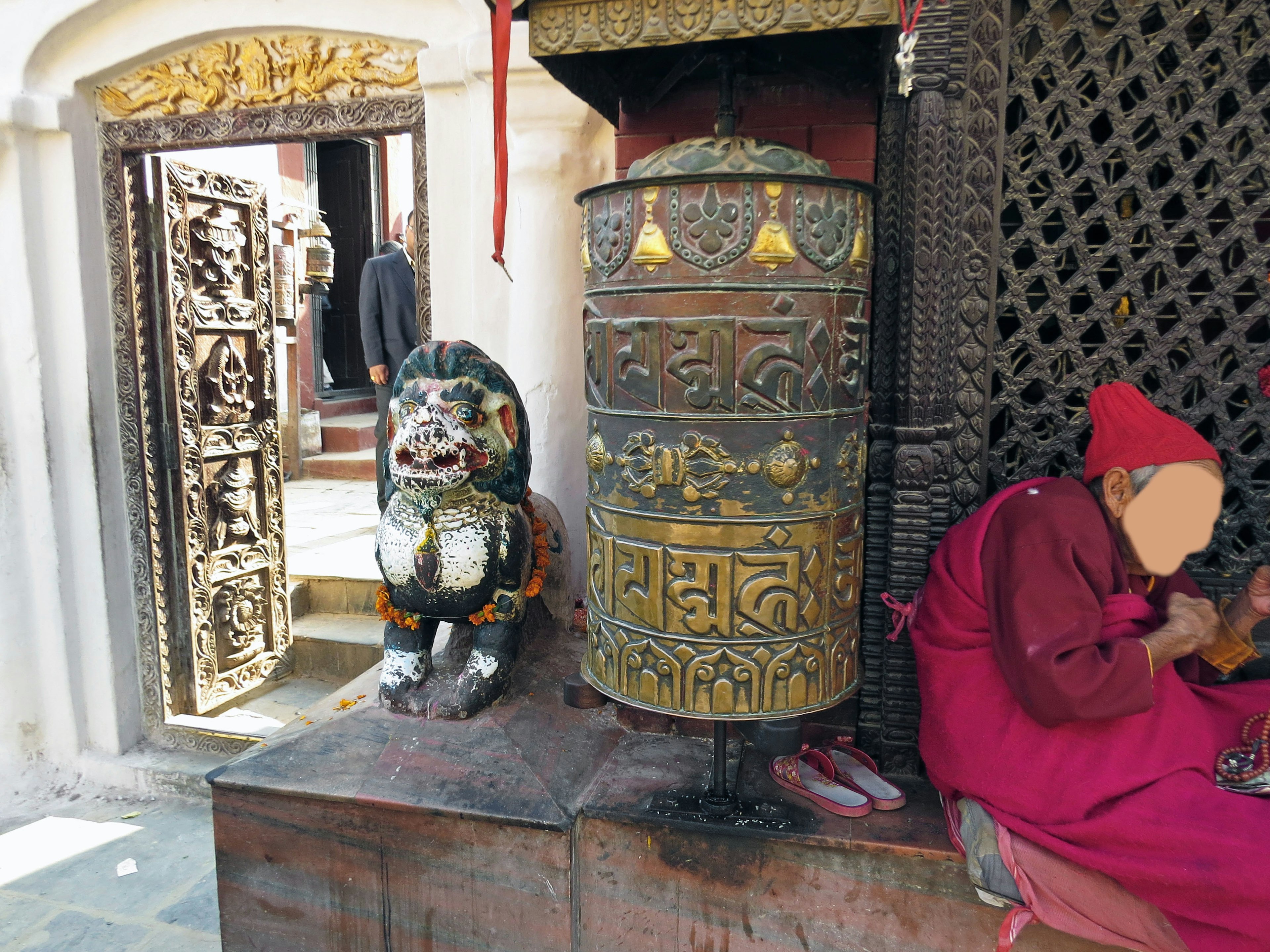 Metal prayer wheel beside a lion statue at a temple entrance with a monk