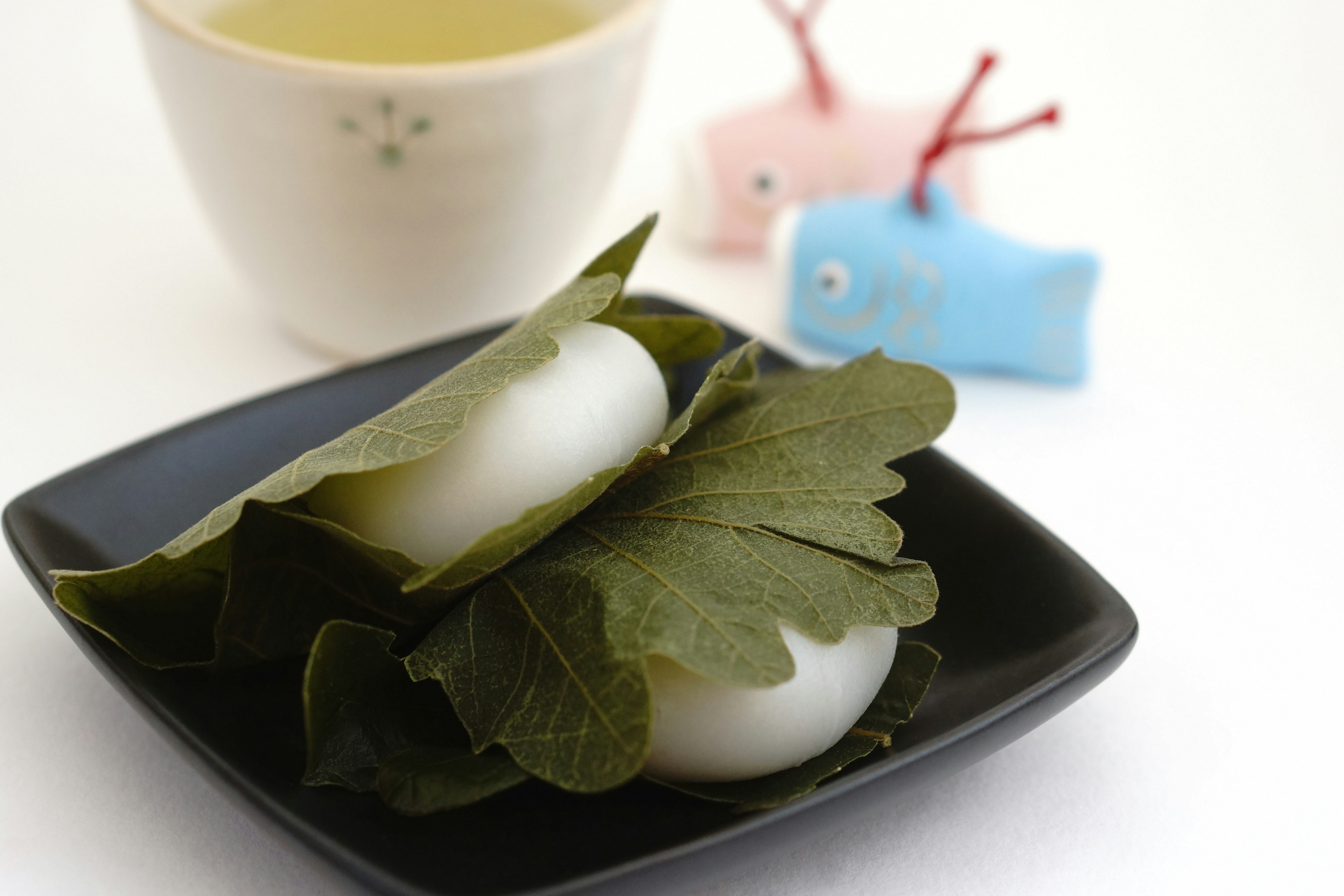 Plate with white rice cakes wrapped in leaves alongside a tea cup and colorful decorative items