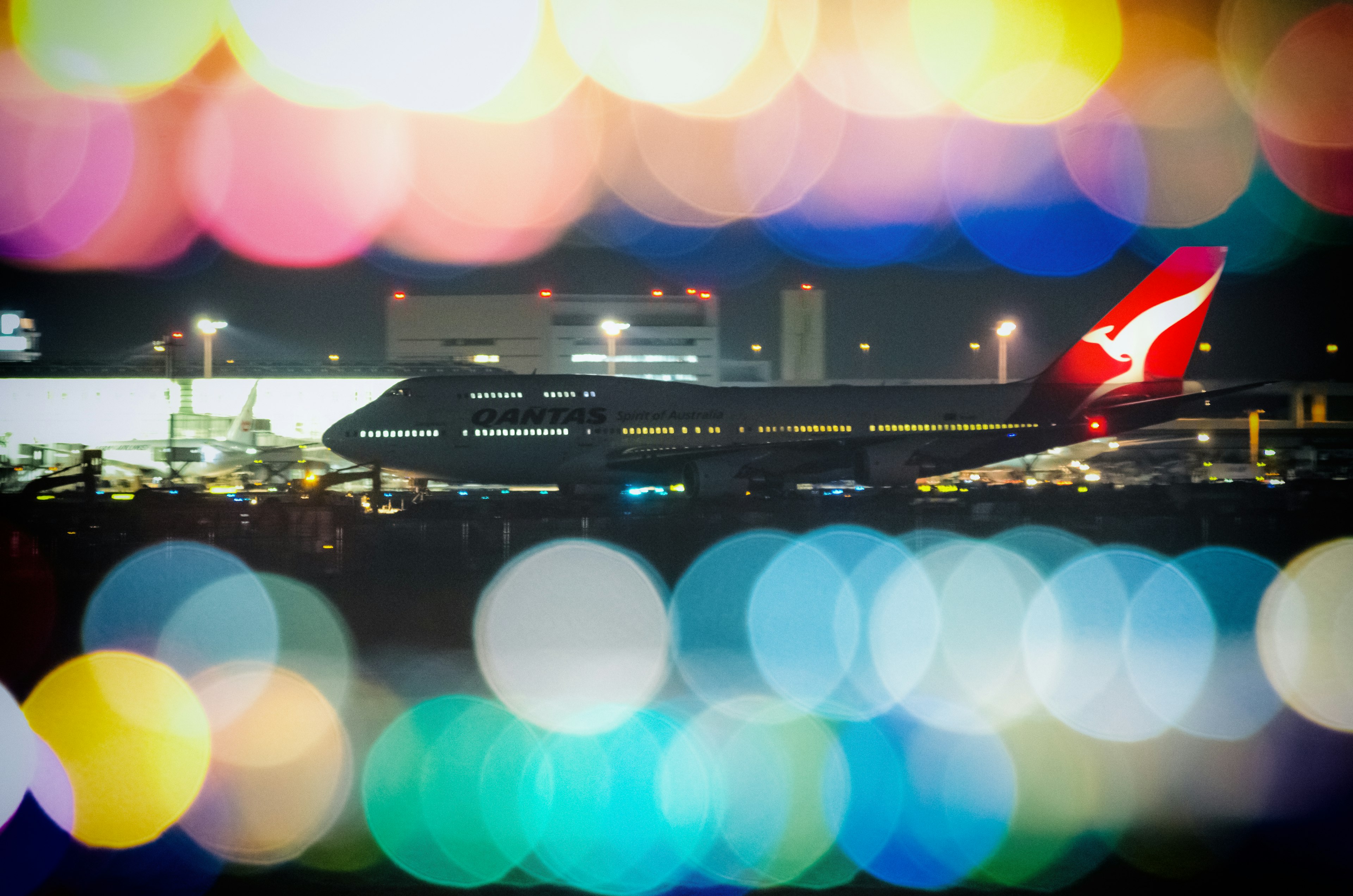 เครื่องบิน Qantas ที่มีเงาอยู่ในพื้นหลัง bokeh ที่มีสีสันในเวลากลางคืน