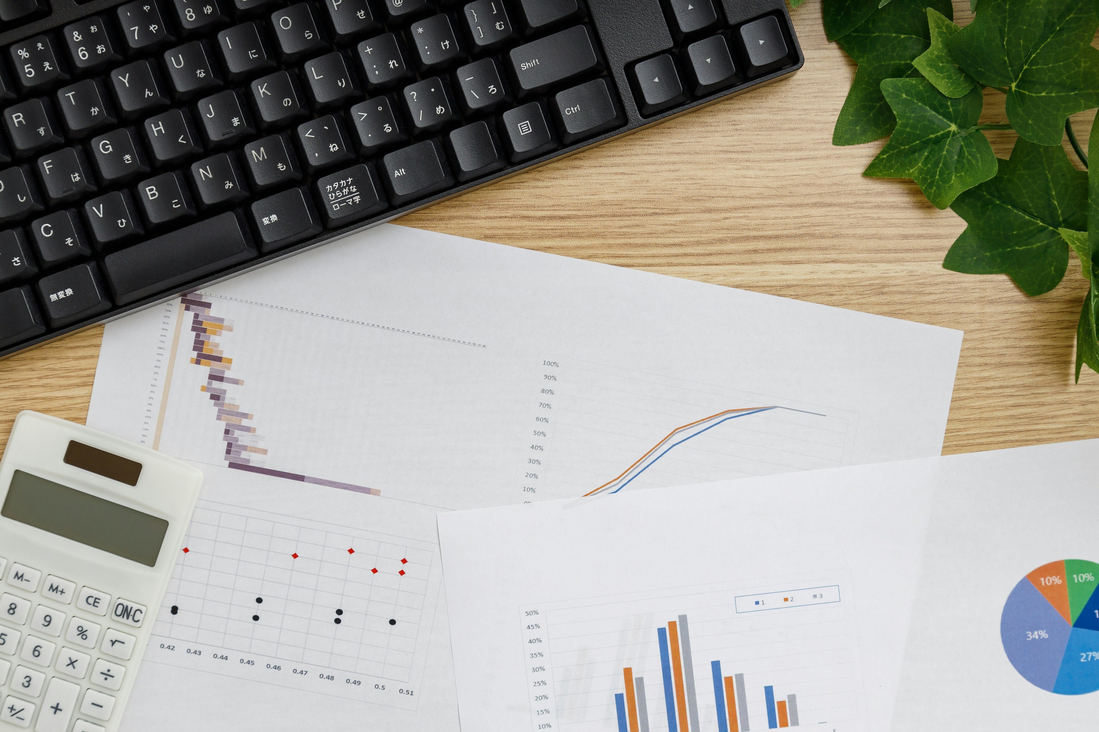 Black keyboard near green plant with graphs and data sheets on the desk