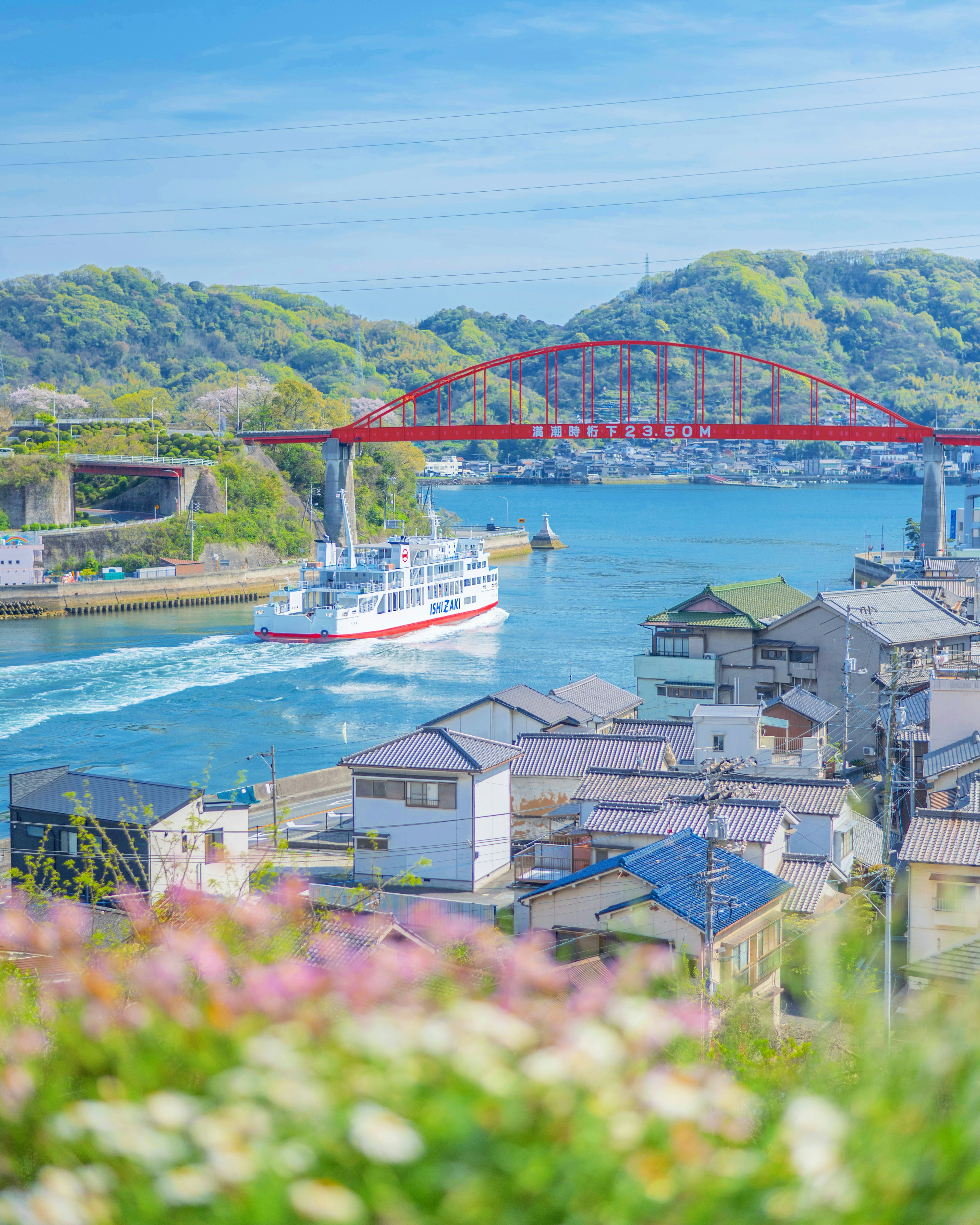 Scenic view of a river under a blue sky featuring a red bridge and a passing boat with houses lining the shore