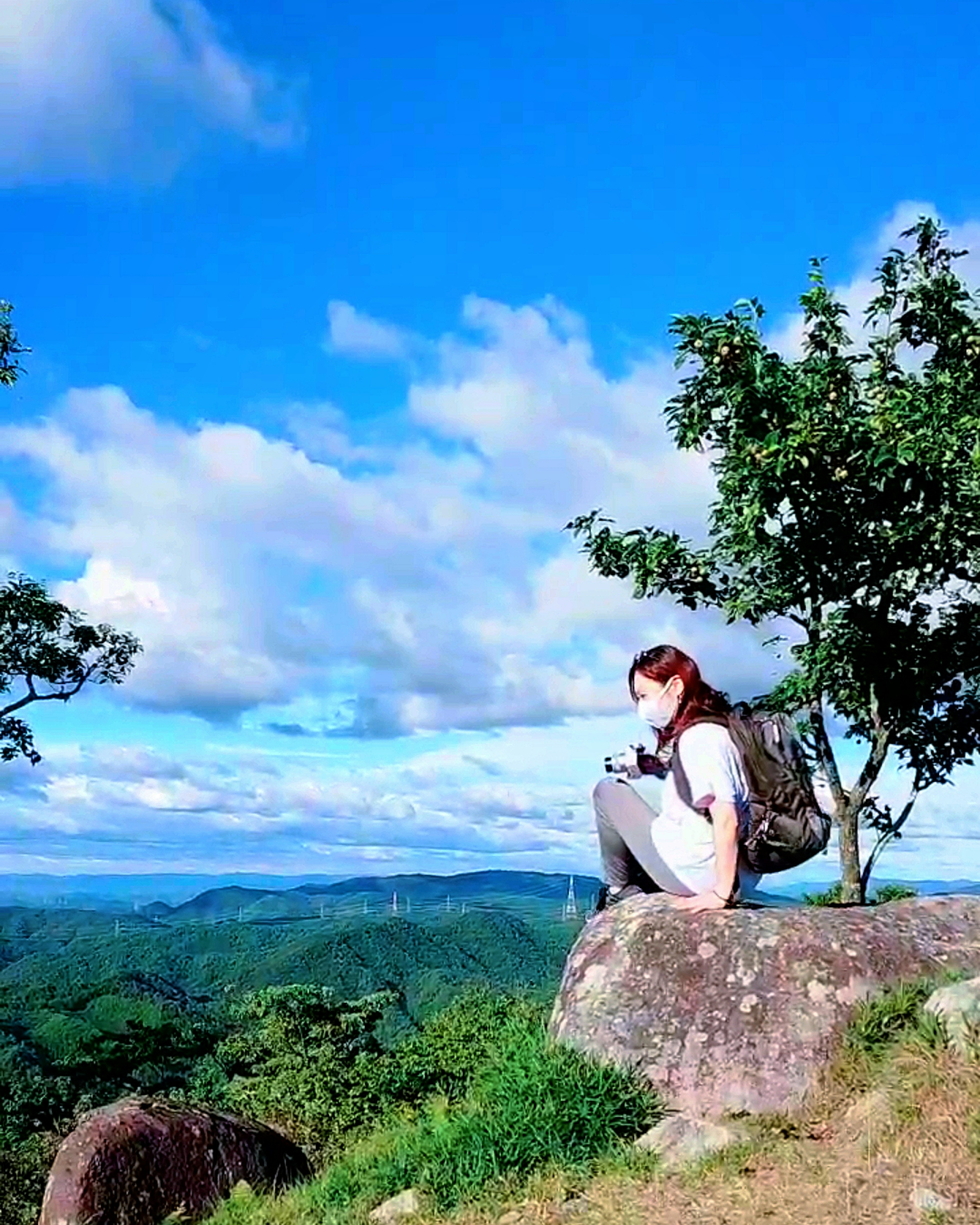 Woman sitting on a rock under a blue sky with clouds and green landscape