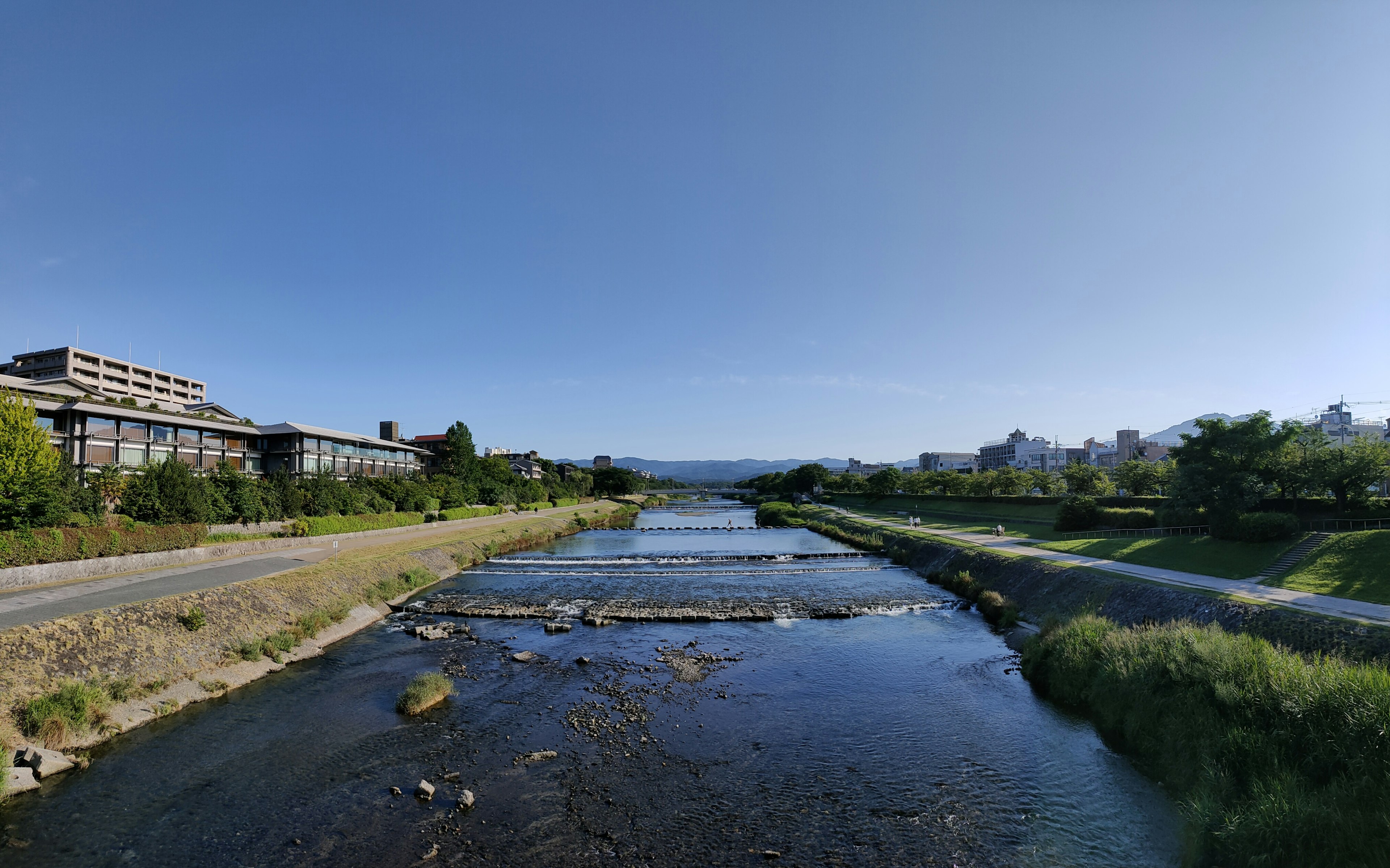 A landscape featuring a river under a blue sky with greenery and distant mountains