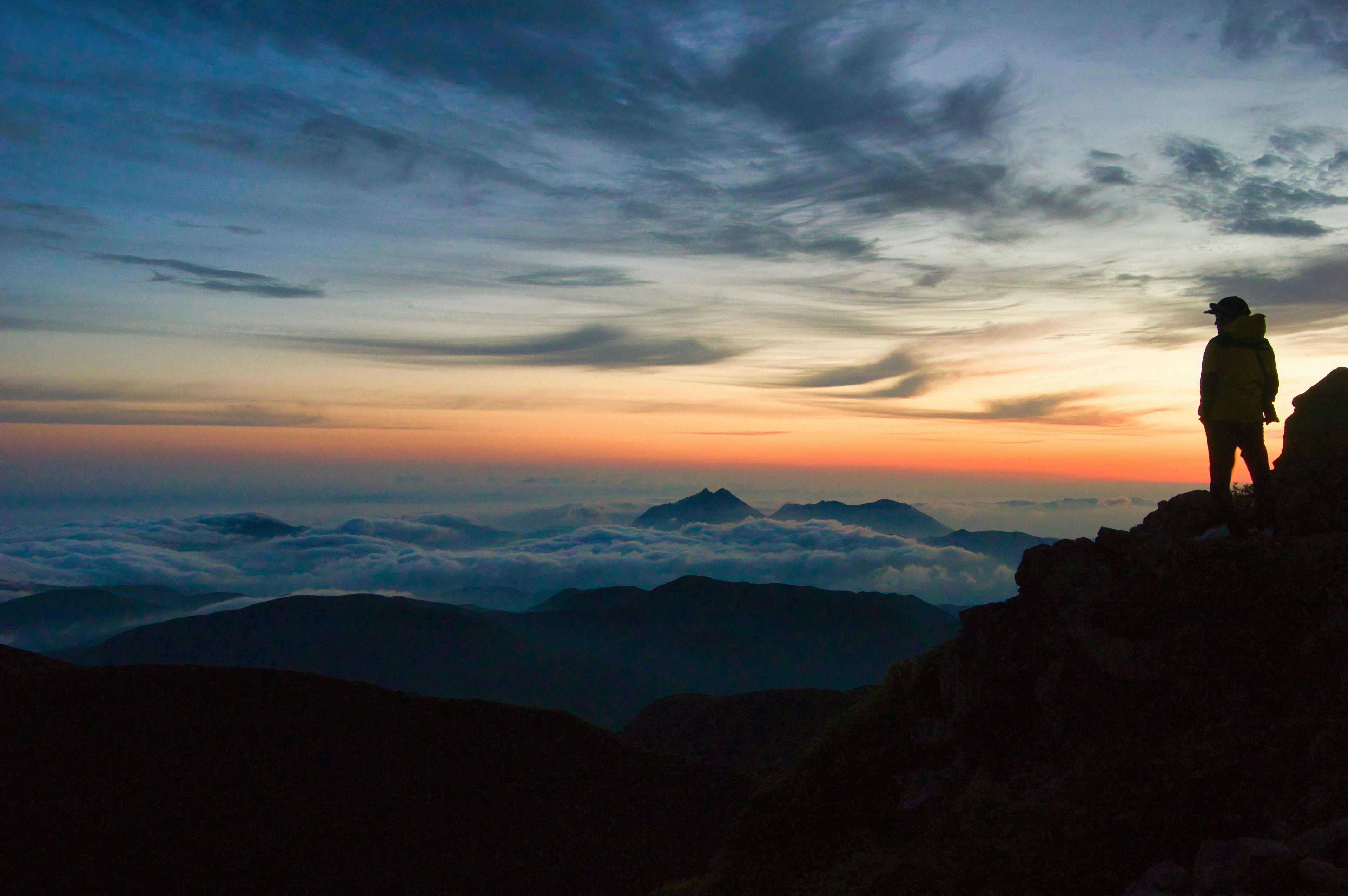 Persona in piedi su una cima montuosa al tramonto con un mare di nuvole