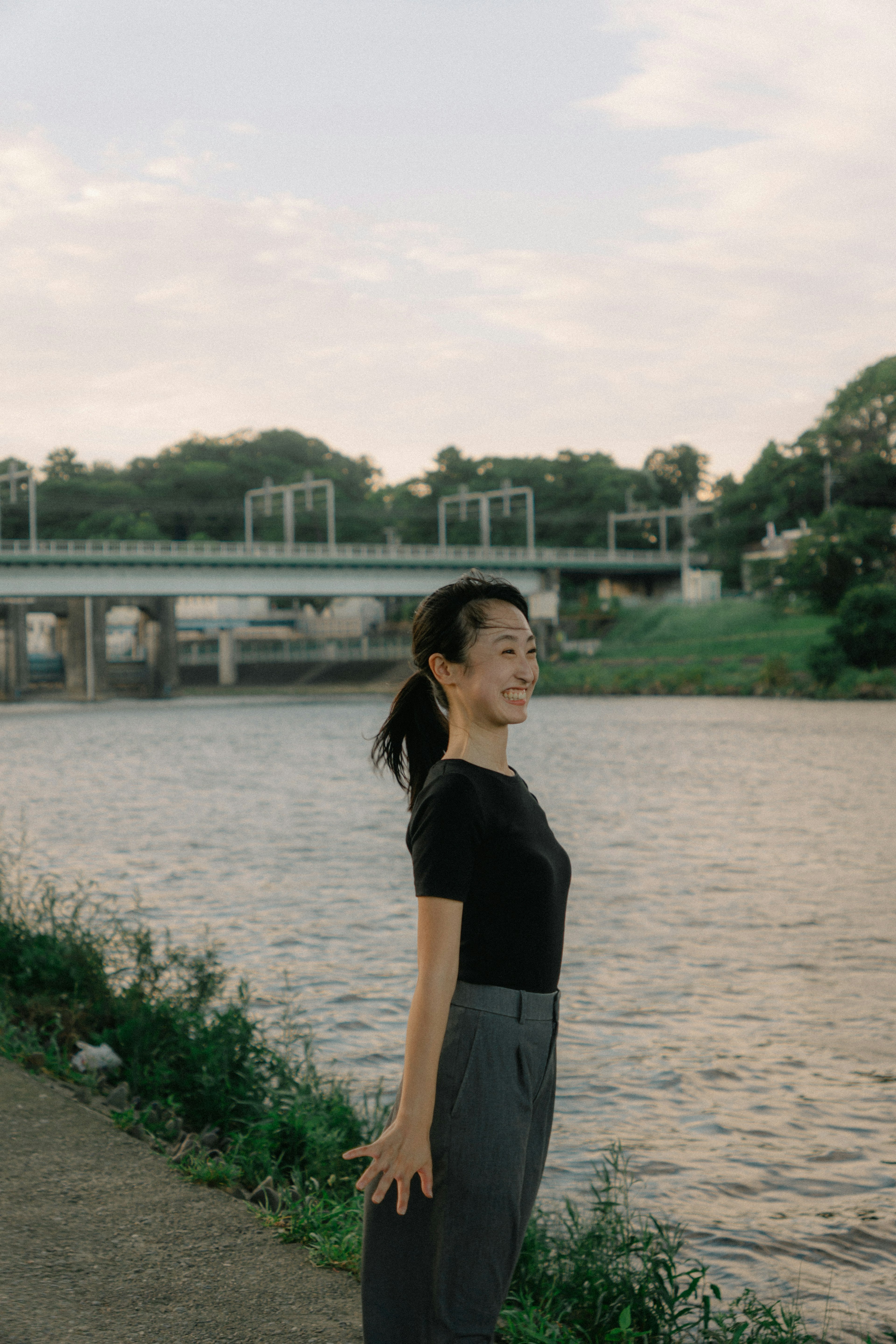 川のそばでポーズをとる女性の横顔　黒のトップスとグレーのパンツ　穏やかな風景