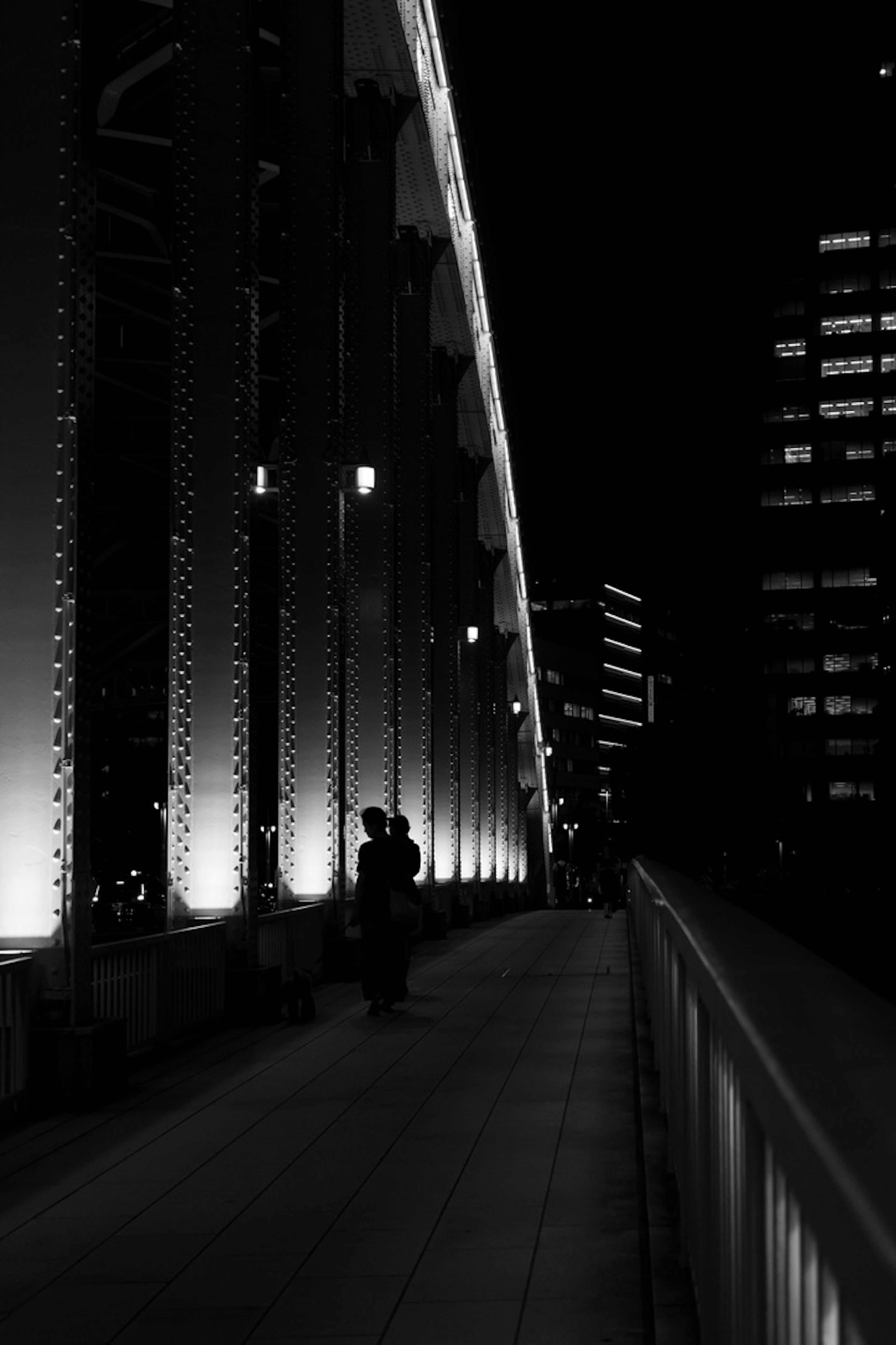 Monochrome photo of a person walking on a bridge at night with striking lights