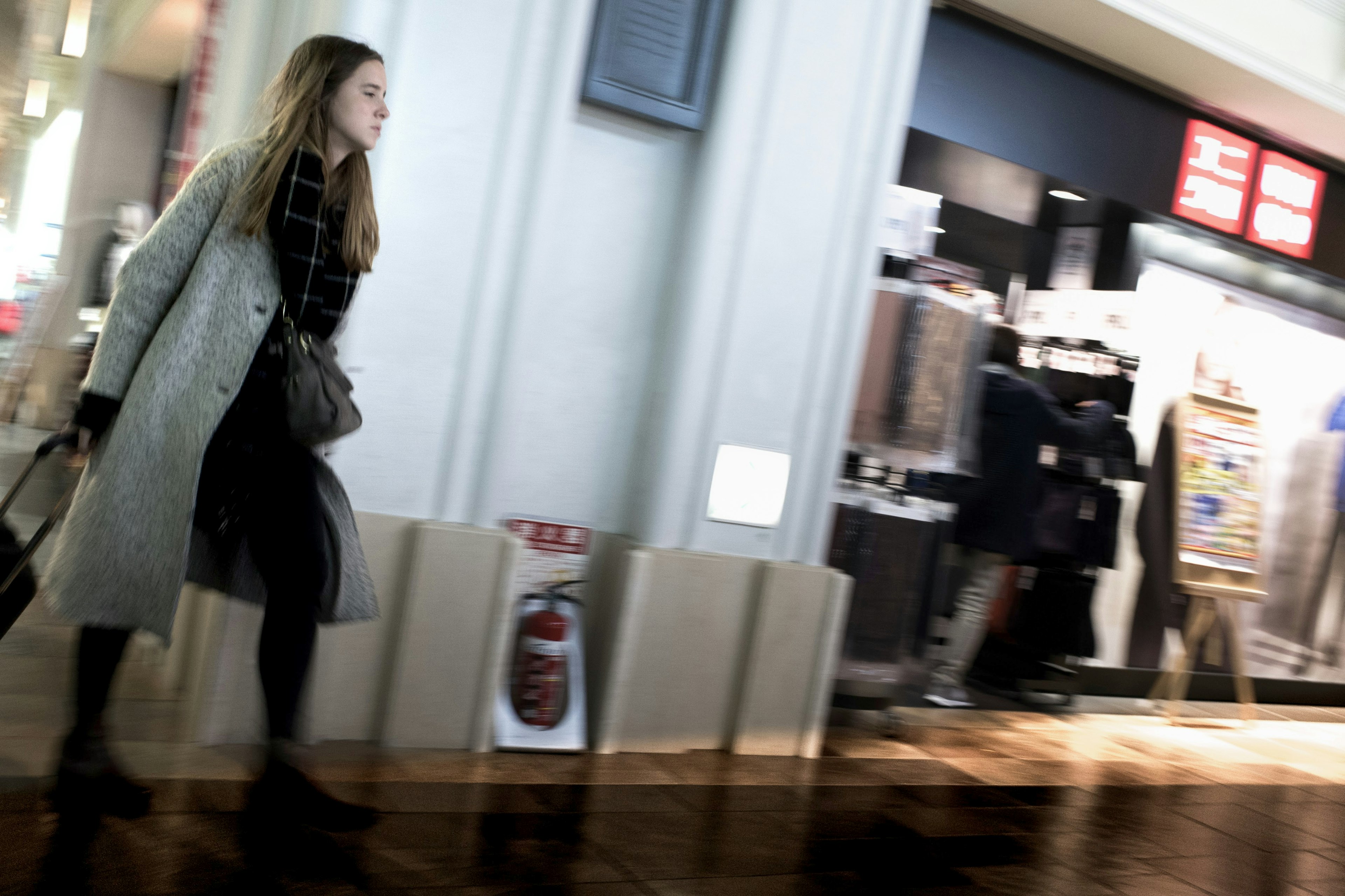 A woman walking in a shopping mall wearing a coat and pulling a cart