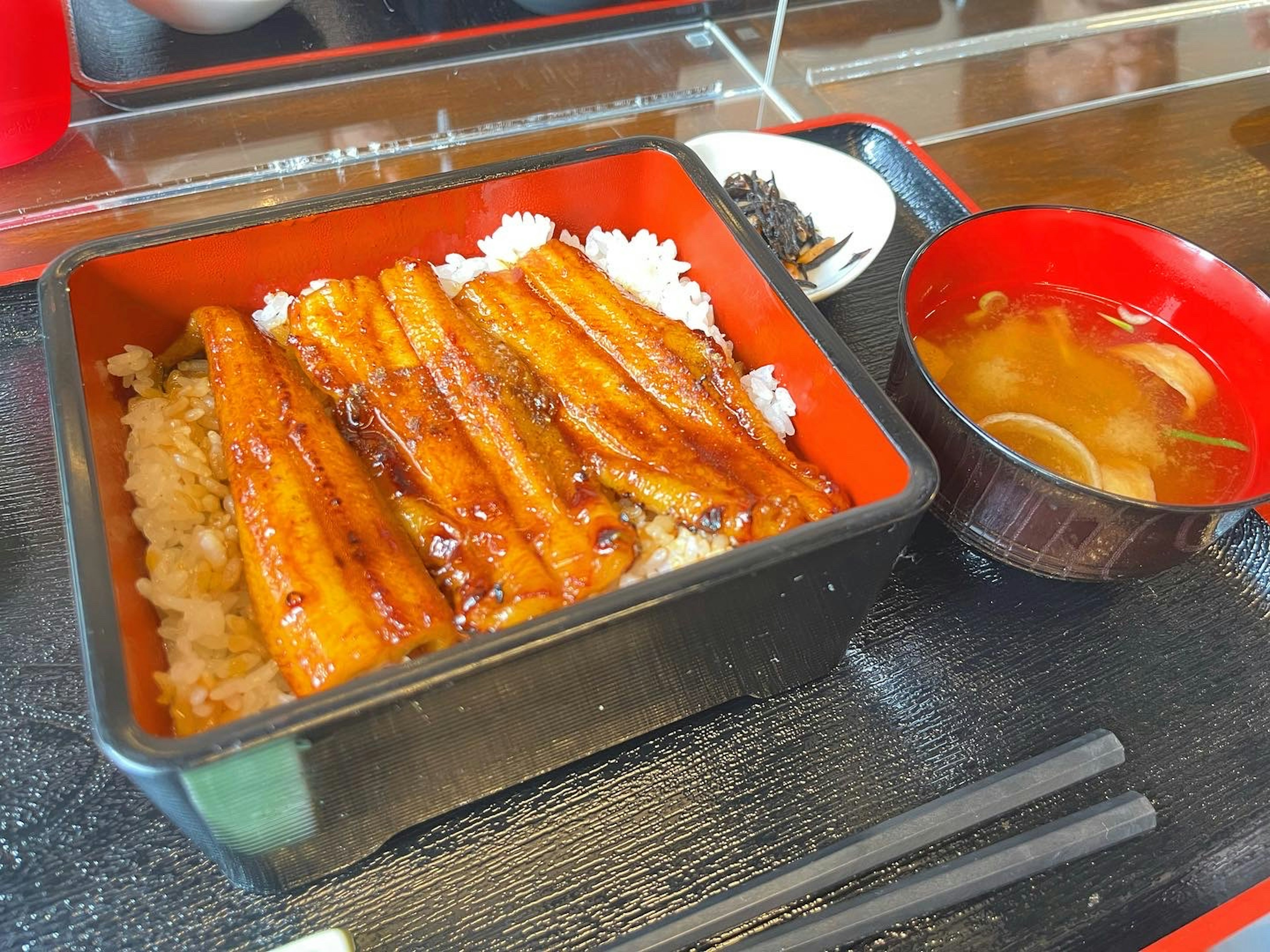 맛있는 장어 덮밥이 빨간 도시락에 담겨 밥과 소스에 유리된 장어가 있다