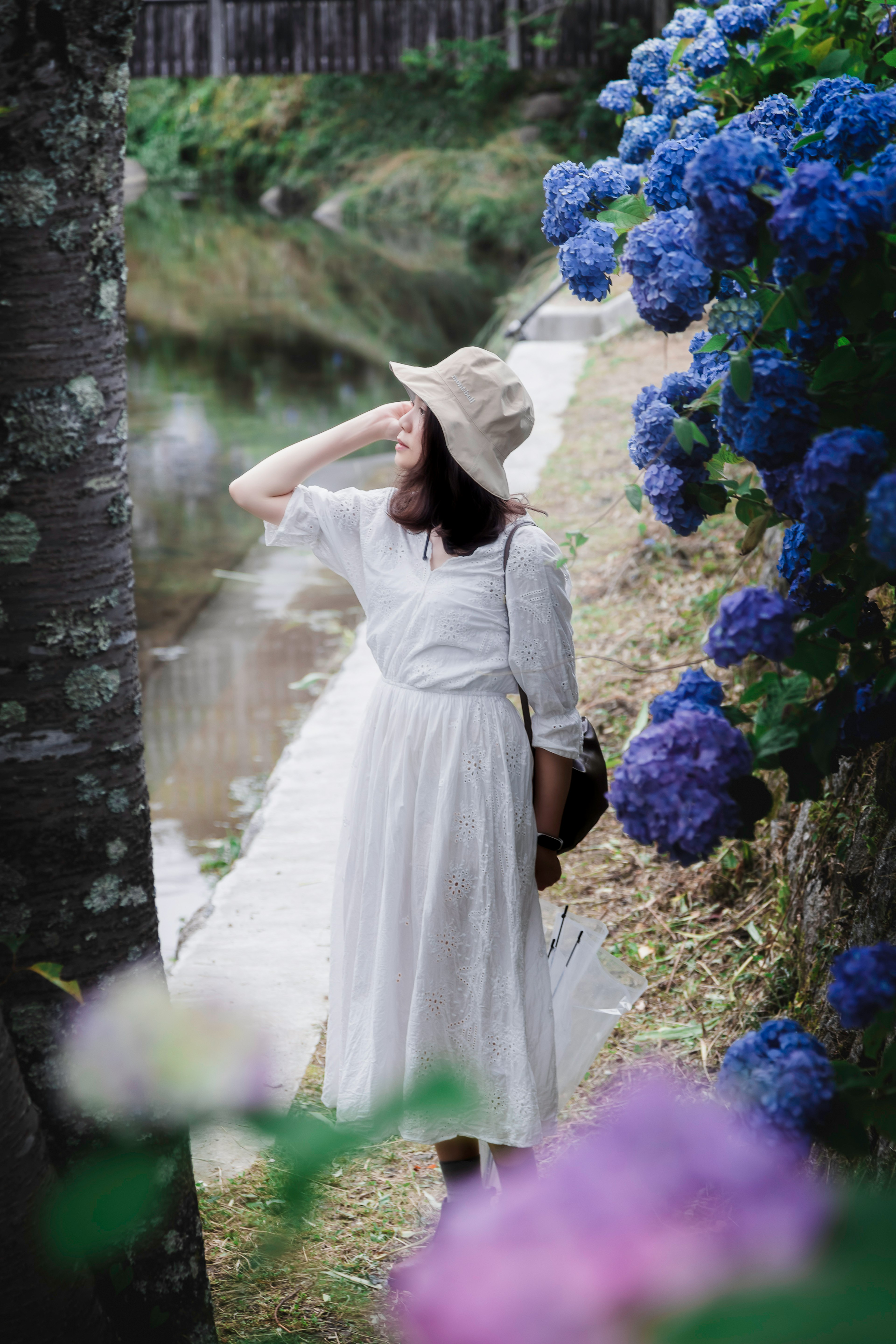 Eine Frau in einem weißen Kleid steht neben blauen Hortensien an einem Fluss