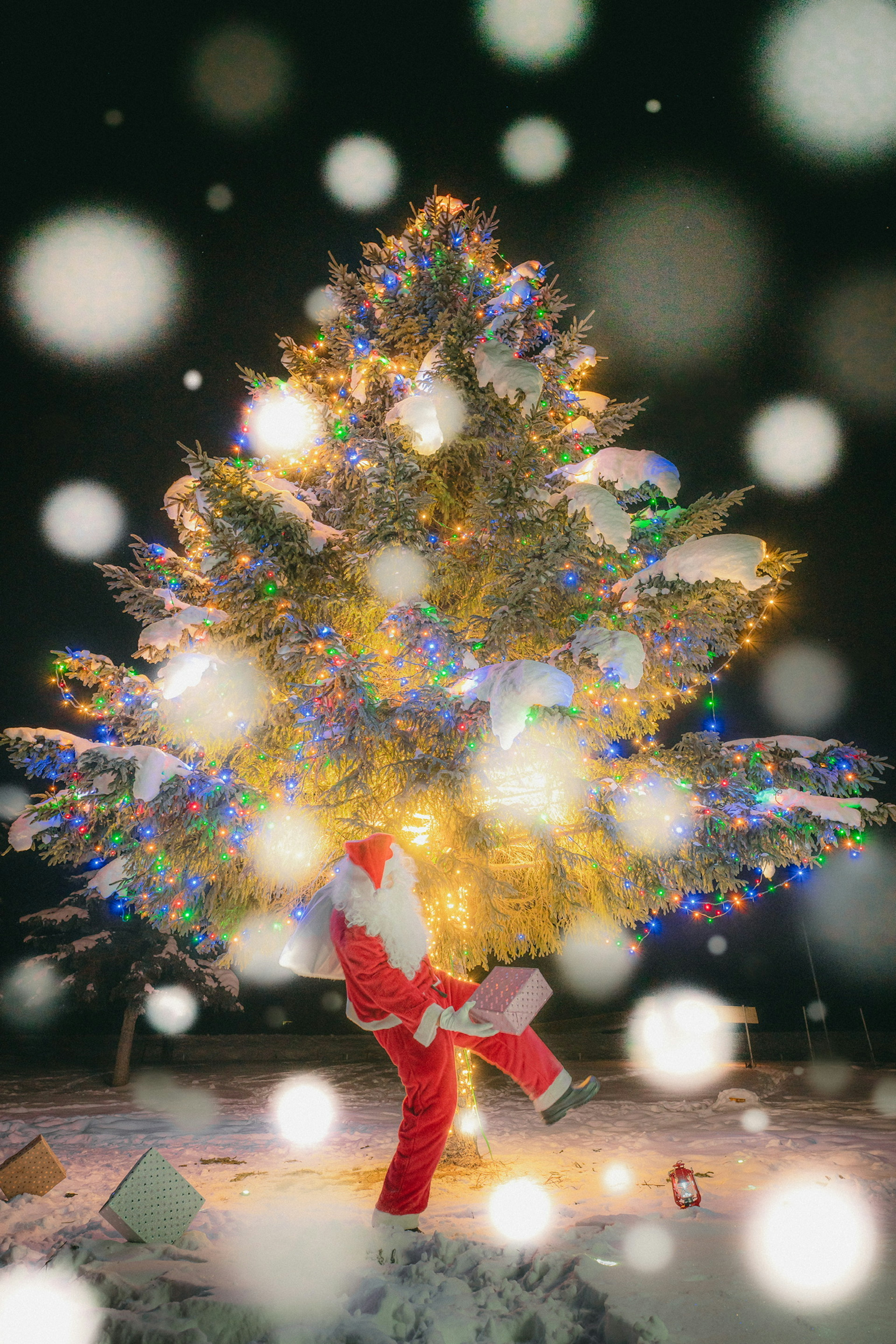 Santa Claus standing in snow with a brightly decorated Christmas tree